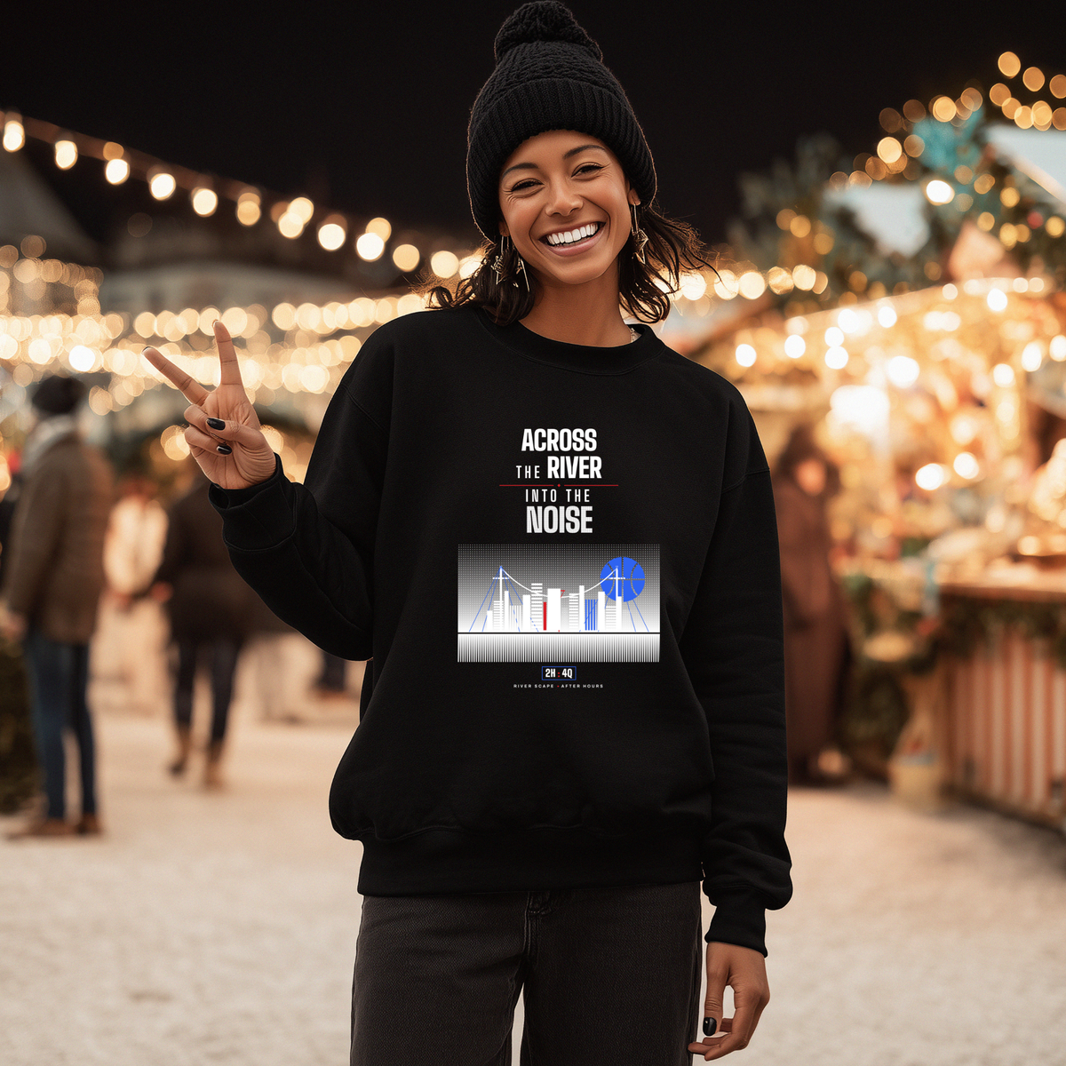 A black woman wearing a black crewneck sweatshirt with a graphic print that reads 'ACROSS THE RIVER INTO THE NOISE',  photographed at a festive outdoor night market with warm string lights in the background.