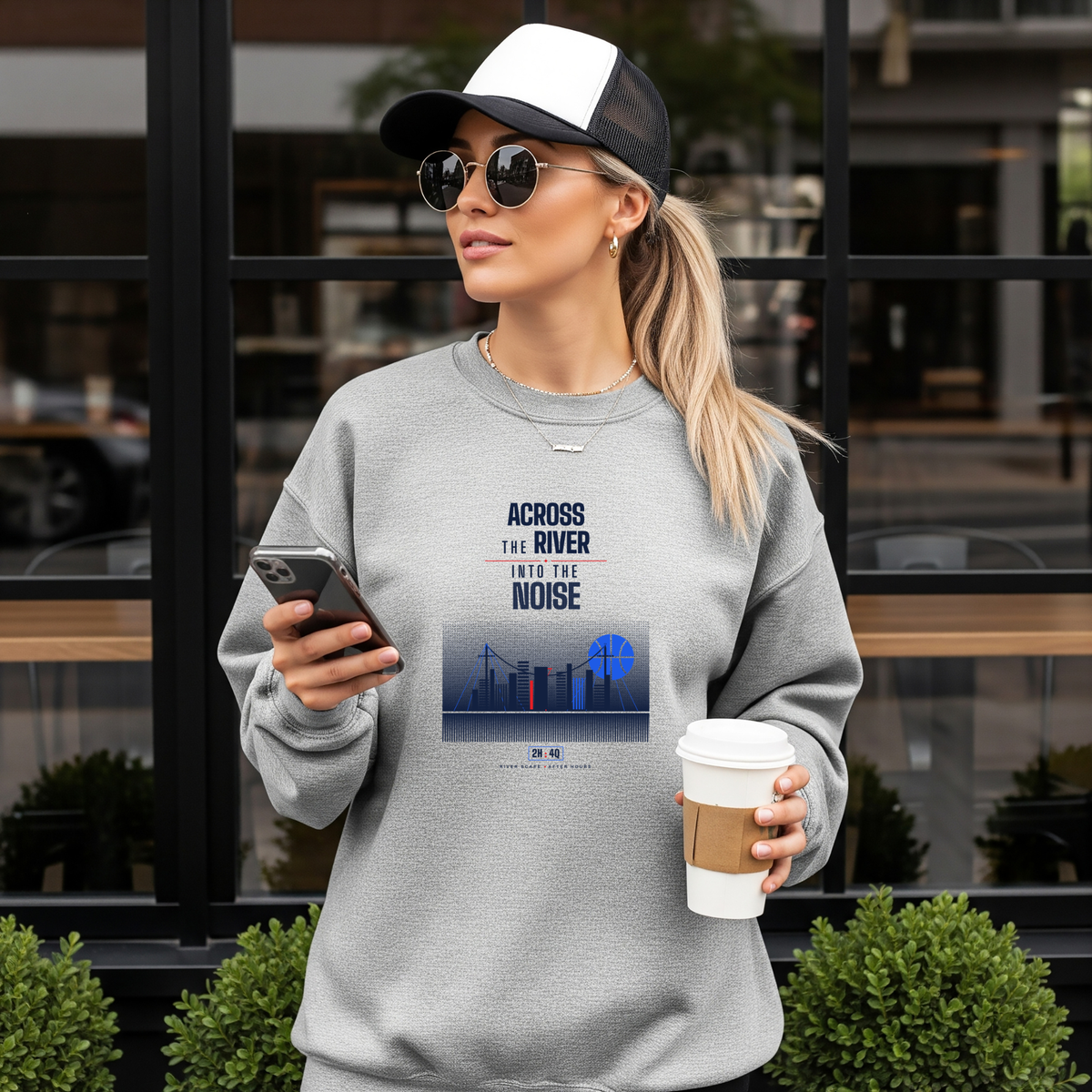 A woman wearing a light gray crewneck sweatshirt with a graphic print that reads 'ACROSS THE RIVER INTO THE NOISE' stands in front of a storefront. She is holding a smartphone and a takeaway coffee cup, wearing a black and white trucker hat and sunglasses.