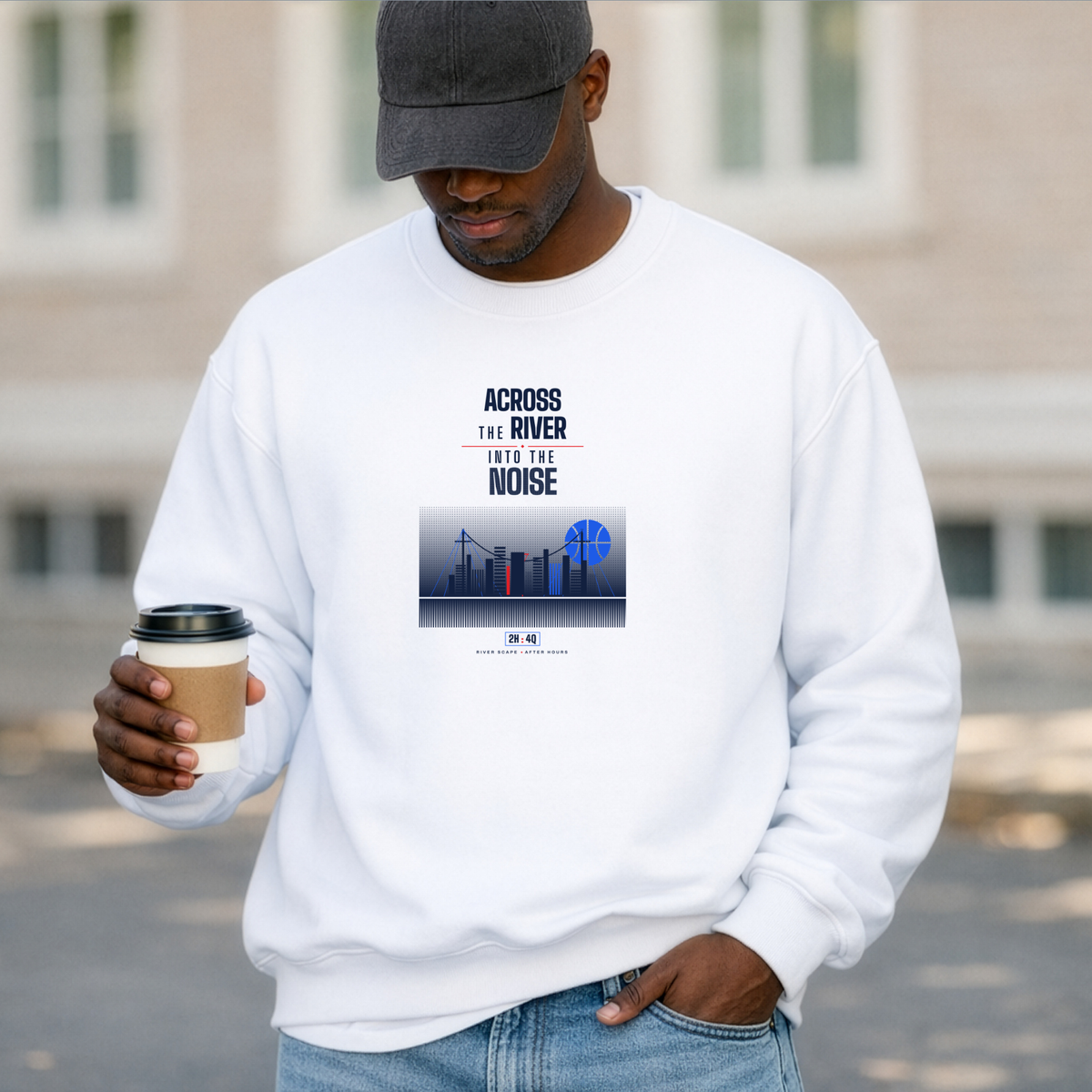 A black man wearing a white crewneck sweatshirt with a graphic print that reads 'ACROSS THE RIVER INTO THE NOISE' above a city skyline and a blue globe. He is wearing a dark gray baseball cap and holding a paper coffee cup in his hand, standing outdoors in front of a blurred building.