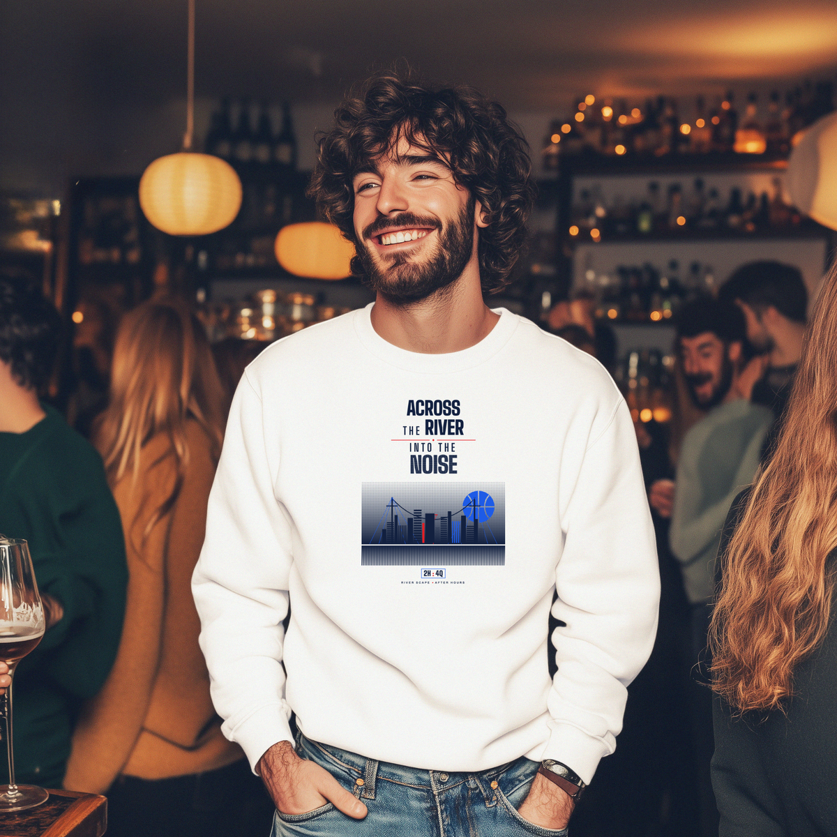 A man smiling in a bar setting wearing a white crewneck sweatshirt with a graphic print that reads 'ACROSS THE RIVER INTO THE NOISE' above a city skyline illustration with a blue basketball shaped moon.
