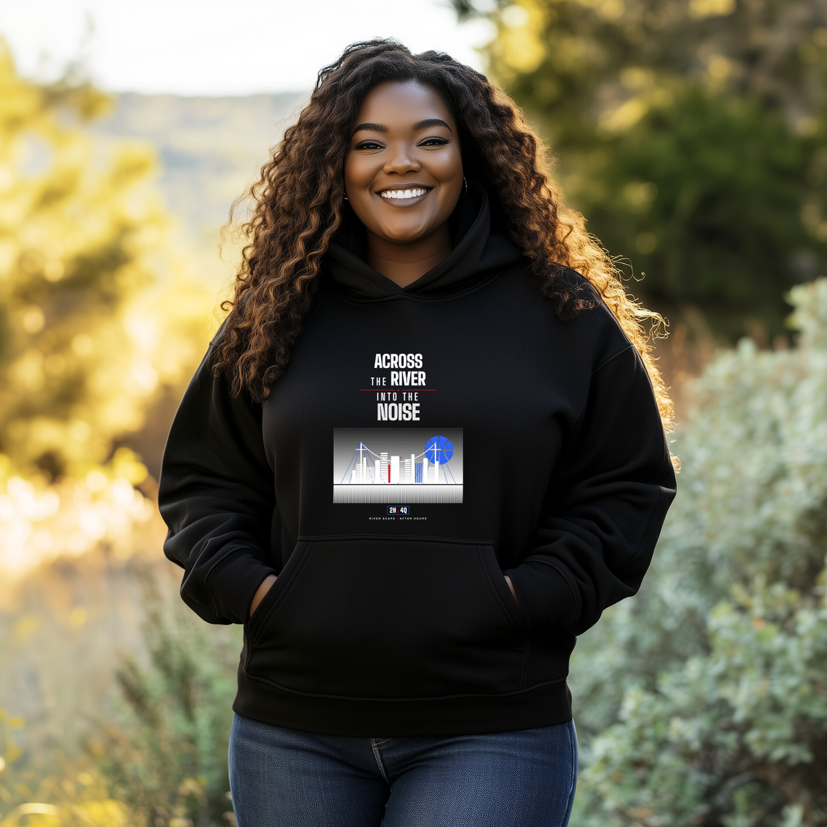 A smiling black woman stands outdoors wearing a black hoodie with a graphic print that reads 'ACROSS THE RIVER INTO THE NOISE'. The background shows blurred greenery and warm sunlight.