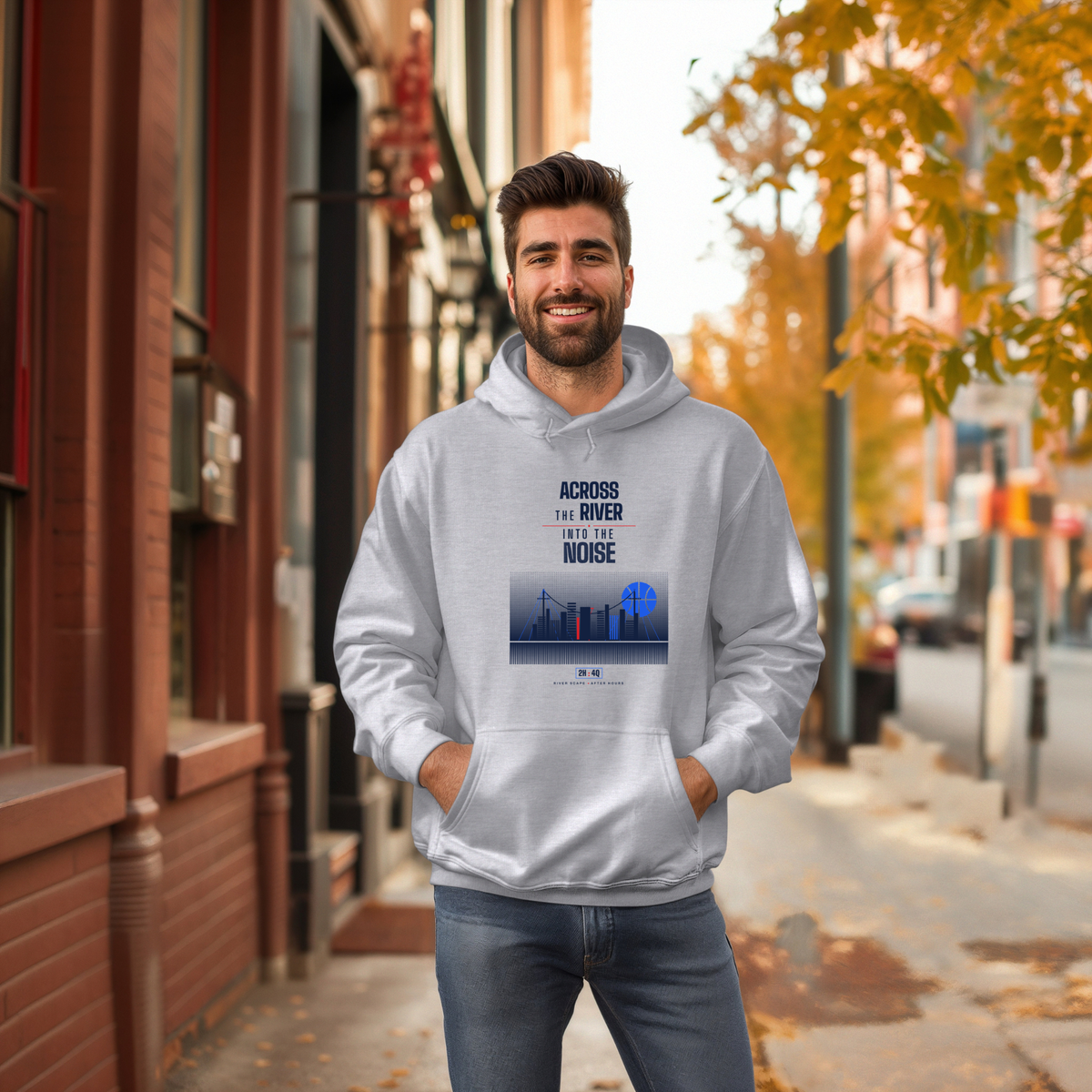 A man wearing a light gray hoodie with a graphic print that reads 'ACROSS THE RIVER INTO THE NOISE' stands on a city sidewalk with autumn trees and brick buildings in the background.