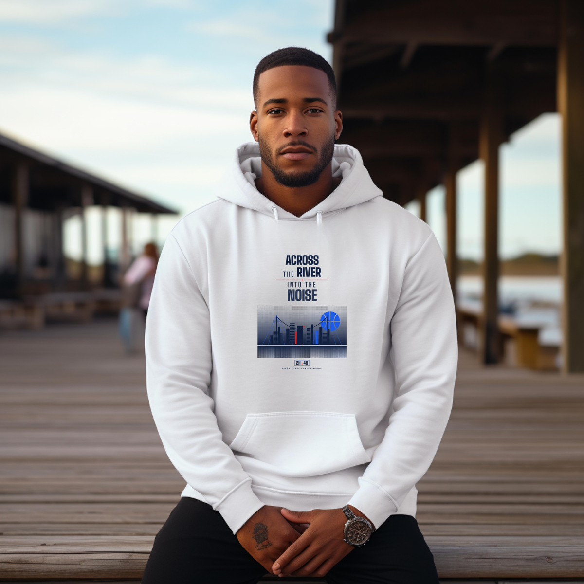 A black man wearing a white pullover hoodie with a blue and black graphic print on the chest, sitting on a wooden boardwalk. The hoodie features the text 'ACROSS THE RIVER INTO THE NOISE'  graphic.