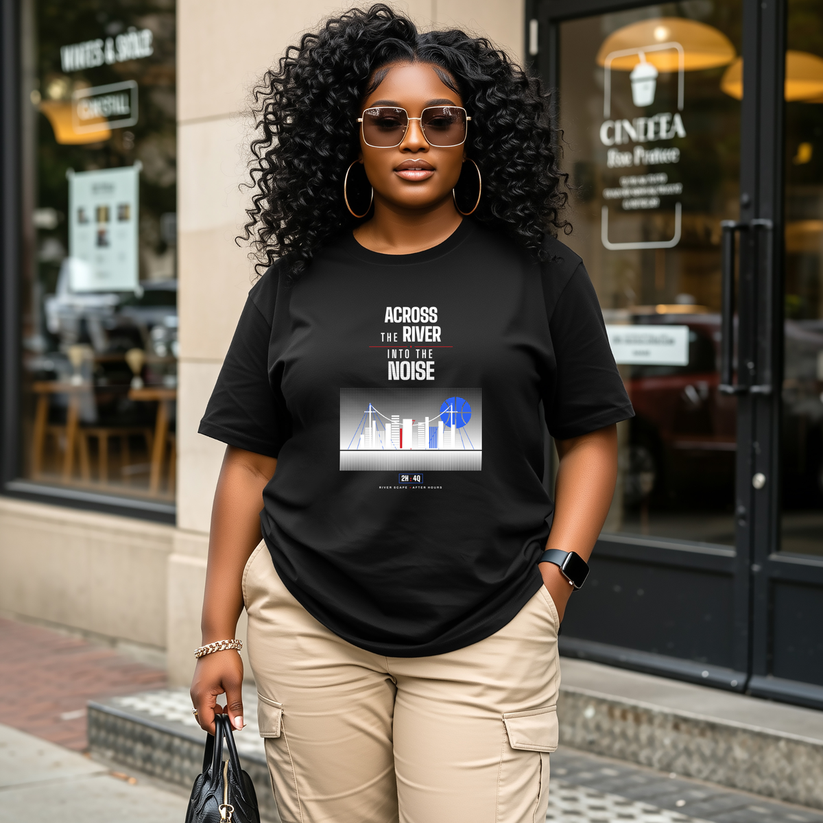 A black woman wearing a black graphic t-shirt with a white and blue 'Across the River' print, styled with beige cargo pants and sunglasses, standing in front of a storefront.
