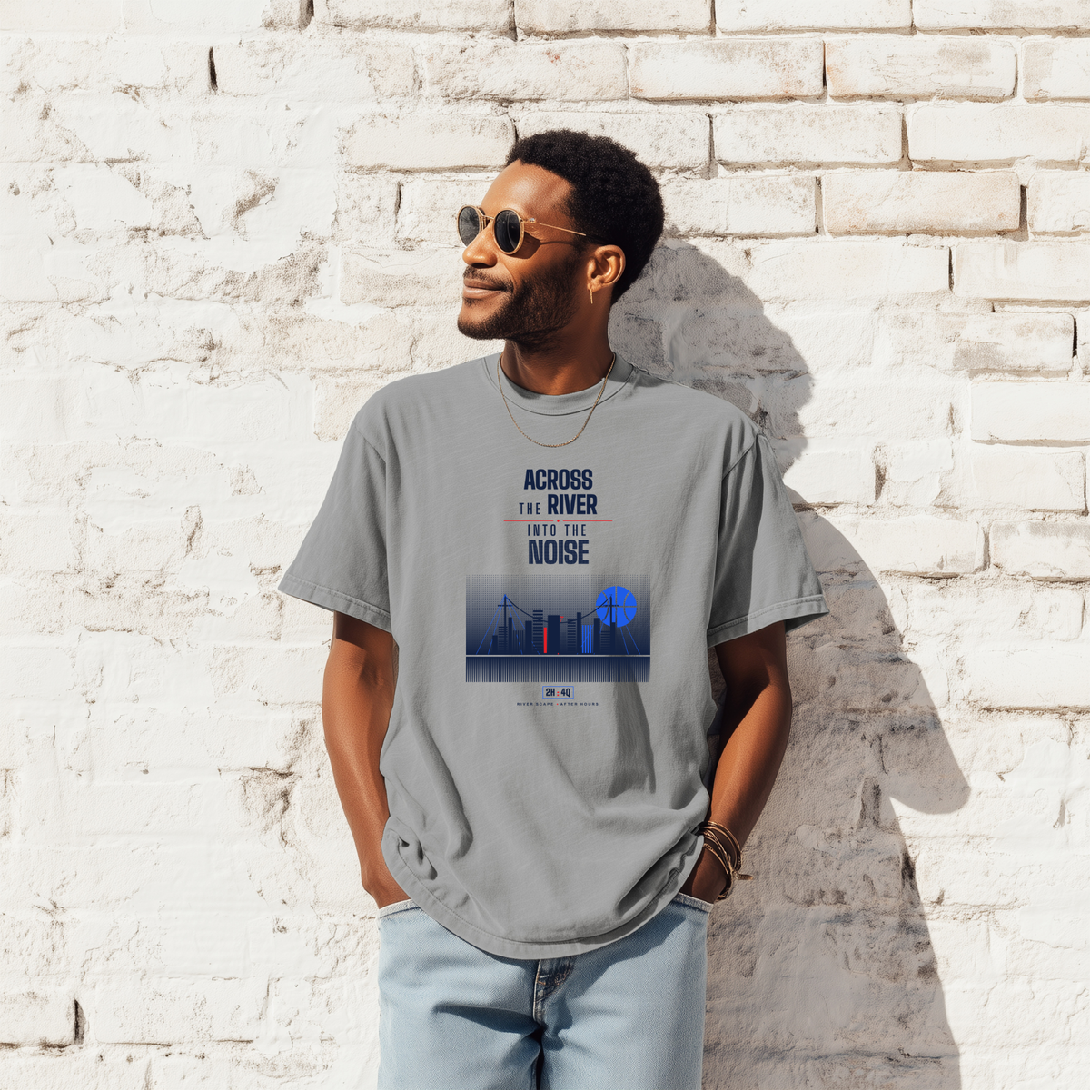 A black man wearing a gray graphic t-shirt with a blue and white print that reads 'ACROSS THE RIVER AND THE NOISE' above an industrial skyline graphic, standing against a white brick wall.