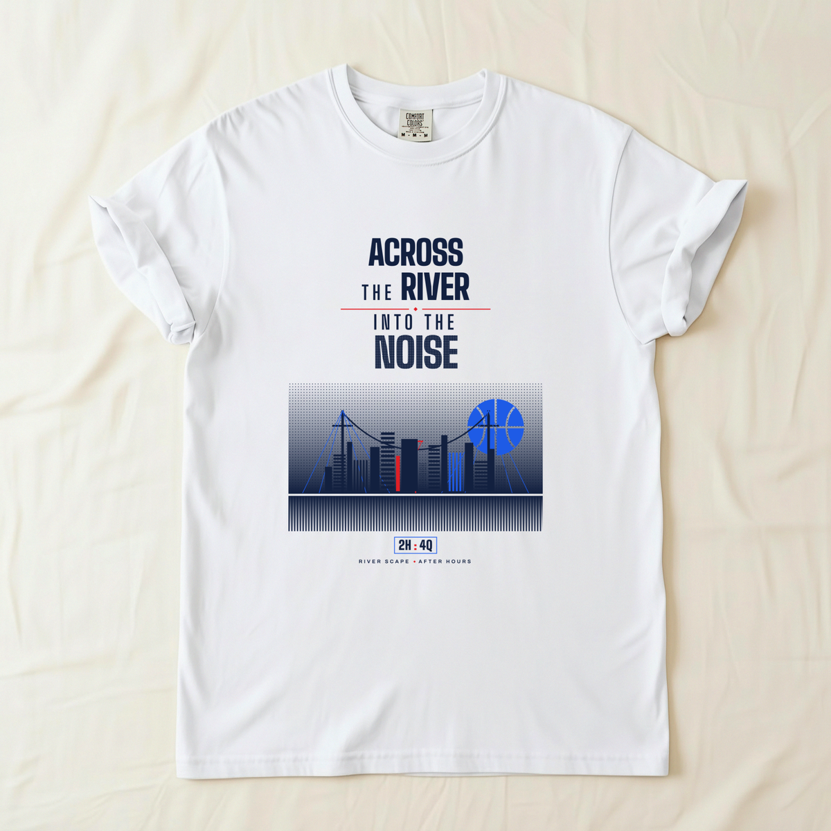A white short-sleeve crewneck t-shirt laid flat on a light beige fabric background. The shirt features a graphic print with the text 'ACROSS THE RIVER INTO THE NOISE' in navy blue and red, above a stylized city skyline illustration with blue basketball shaped moon. 