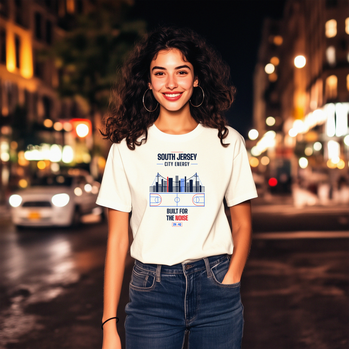A smiling woman with curly dark hair stands on a blurred city street at night wearing a white graphic T-shirt and blue jeans. The T-shirt has a blue and red South Jersey City Energy graphic with the text 'SOUTH JERSEY CITY ENERGY' and 'BUILT FOR THE NOISE'.
