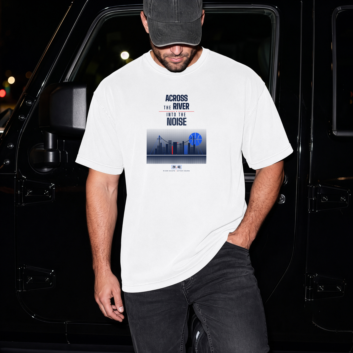 A man wearing a white graphic t-shirt with a blue and black city skyline print and the text 'ACROSS THE RIVER INTO THE NOISE', paired with black jeans and a black cap, standing in front of a black vehicle at night.