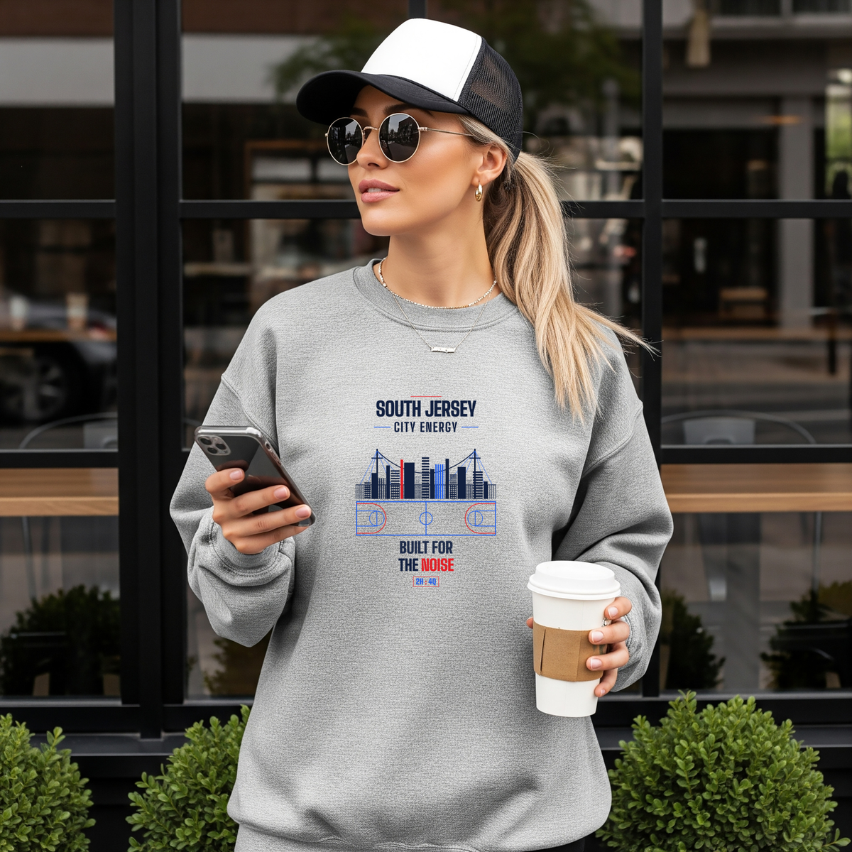 A woman wearing a light gray crewneck sweatshirt with a SOUTH JERSEY CITY ENERGY, BUILT FOR THE NOISE graphic stands outdoors in front of a building with large black-framed windows. She is holding a smartphone and a takeaway coffee cup.