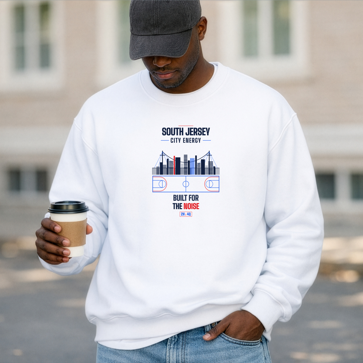 A black man wearing a white crewneck sweatshirt with a SOUTH JERSEY CITY ENERGY, BUILT FOR THE NOISE graphic, paired with a dark gray baseball cap and blue jeans, holding a coffee cup in an outdoor setting.