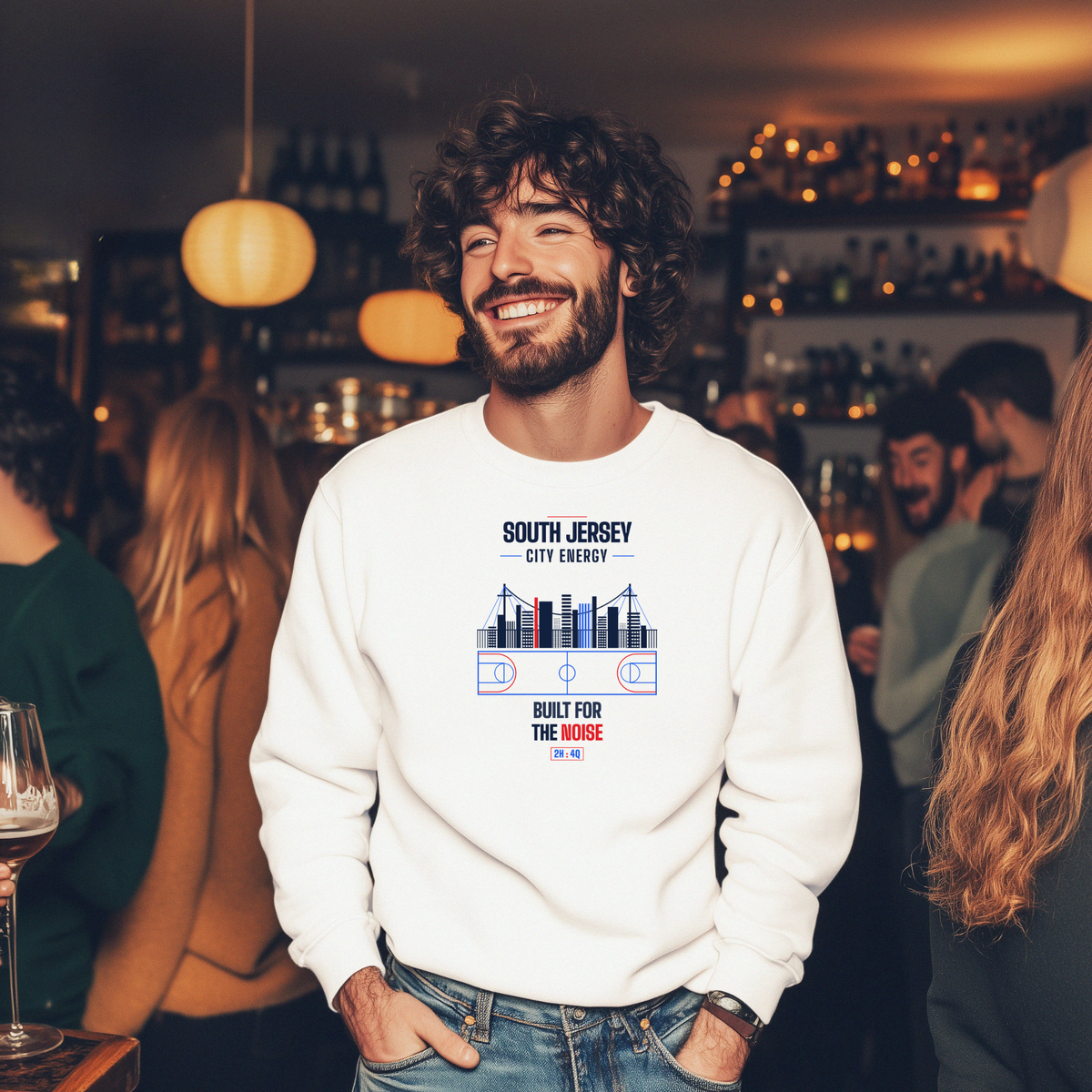 A man smiling in a social bar setting wearing a white crewneck sweatshirt with a graphic print that reads 'SOUTH JERSEY CITY ENERGY' and 'BUILT FOR THE NOISE' with a stylized skyline and sports team logo.