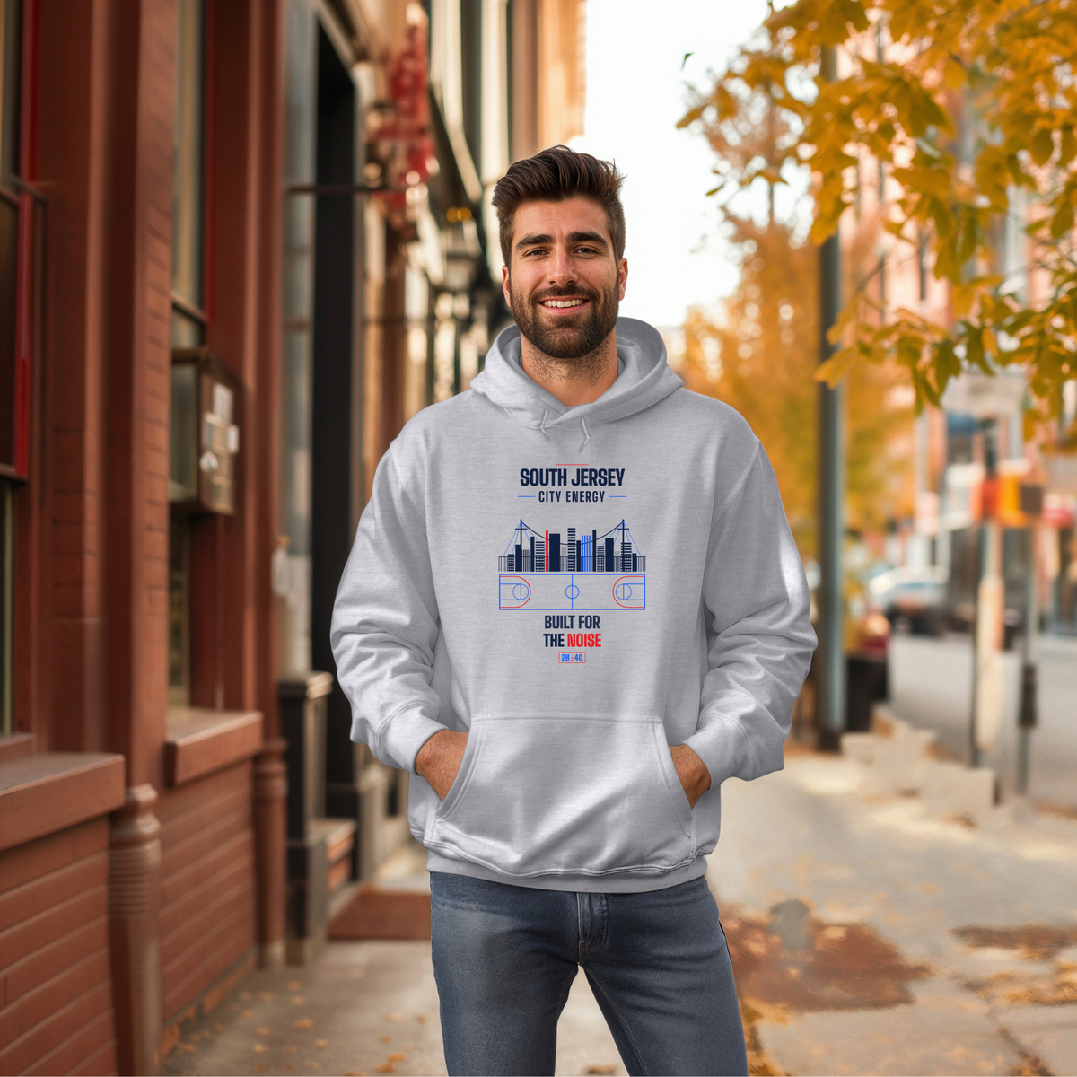 A man wearing a light gray hoodie with a 'South Jersey, City Energy' graphic stands on a city street with brick buildings and autumn trees in the background.