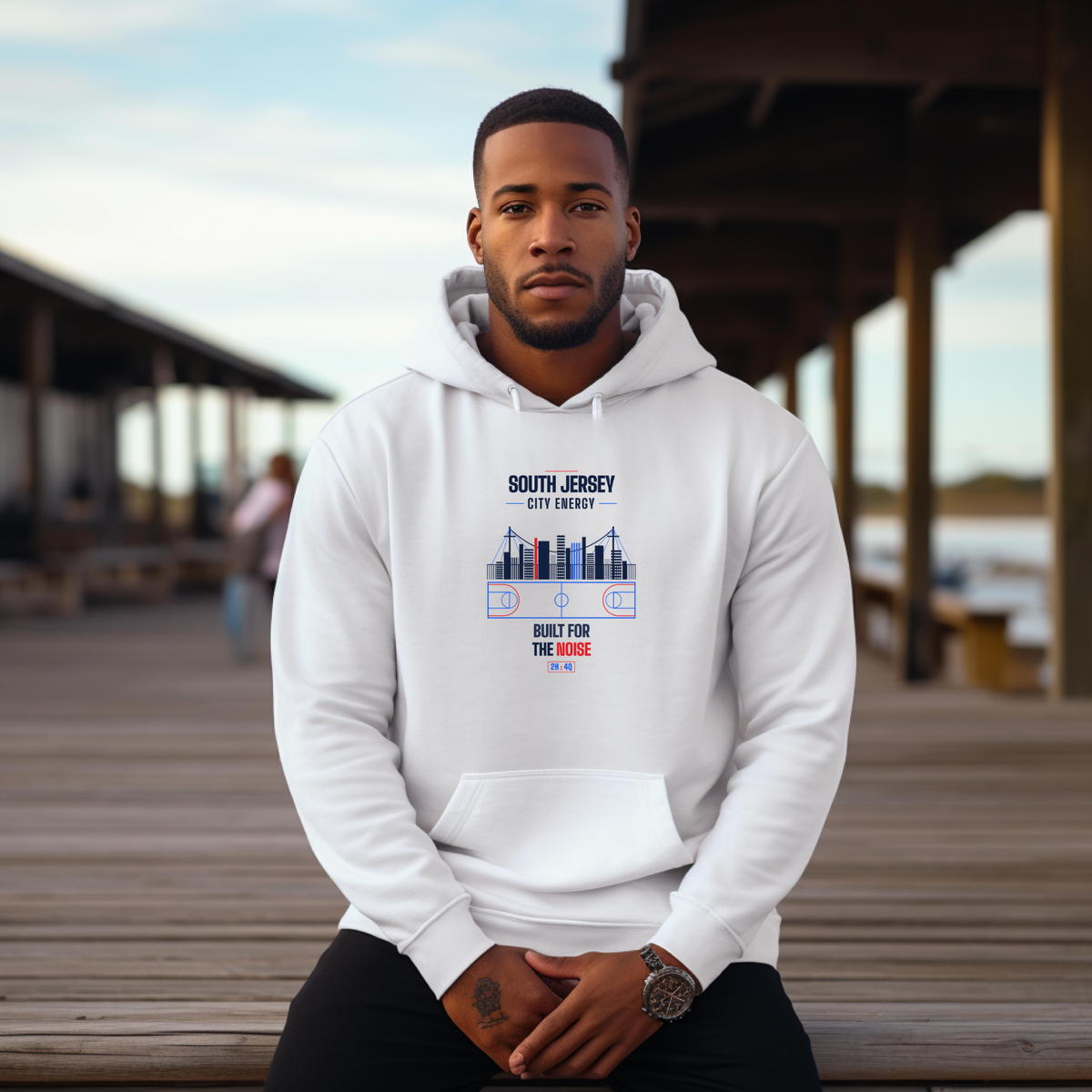 A black man wearing a white hoodie with a South Jersey City, City Energy shore graphic, sitting on a wooden dock in an outdoor setting.