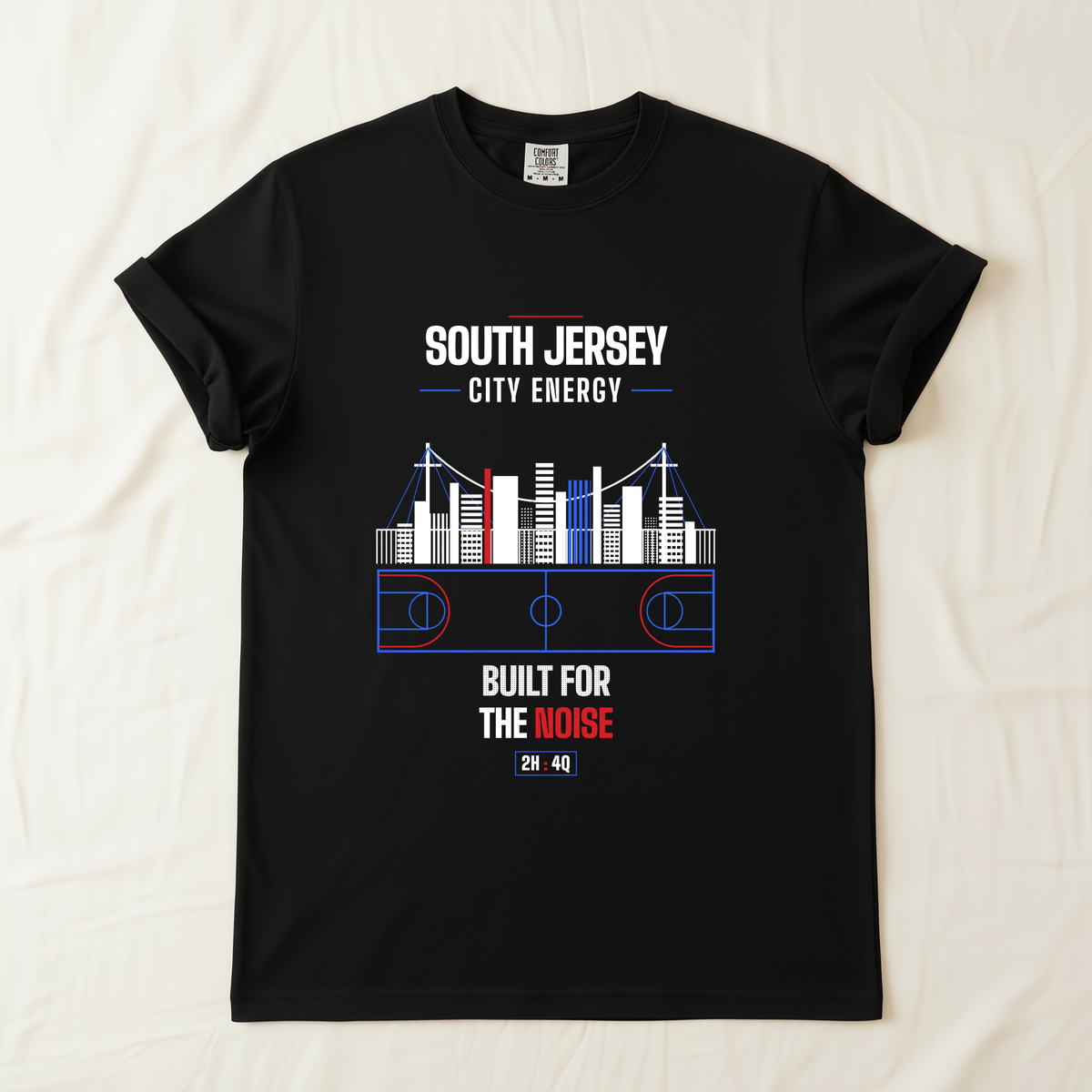 A black short-sleeve t-shirt laid flat on a light beige fabric background. The shirt features a graphic print with the text 'SOUTH JERSEY CITY ENERGY' and 'BUILT FOR THE NOISE' in white and red, surrounding a stylized basketball court and city skyline design in white, blue, and red.