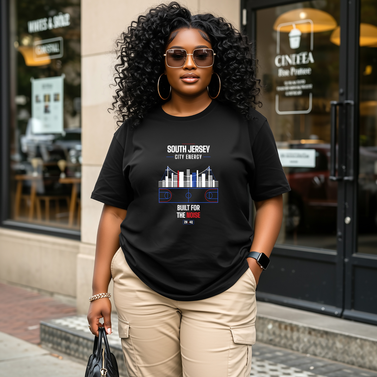 A black woman wearing a black graphic t-shirt with a 'South Jersey City Energy' design stands on a city sidewalk in front of a storefront.
