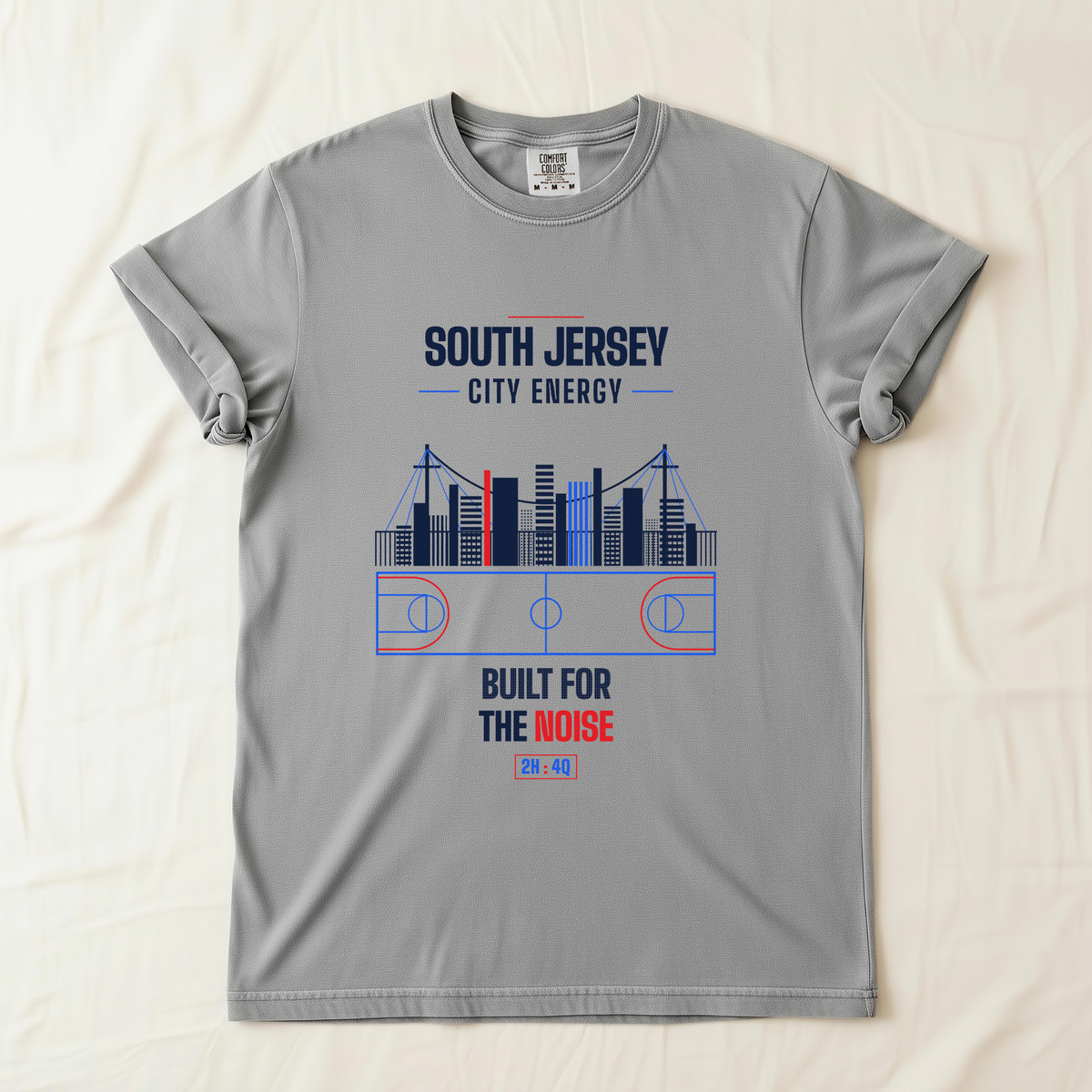 A gray short-sleeve t-shirt laid flat on a light beige fabric background. The shirt features a graphic print of a basketball court and city skyline with the text 'SOUTH JERSEY CITY ENERGY' and 'BUILT FOR THE NOISE' in navy blue and red.