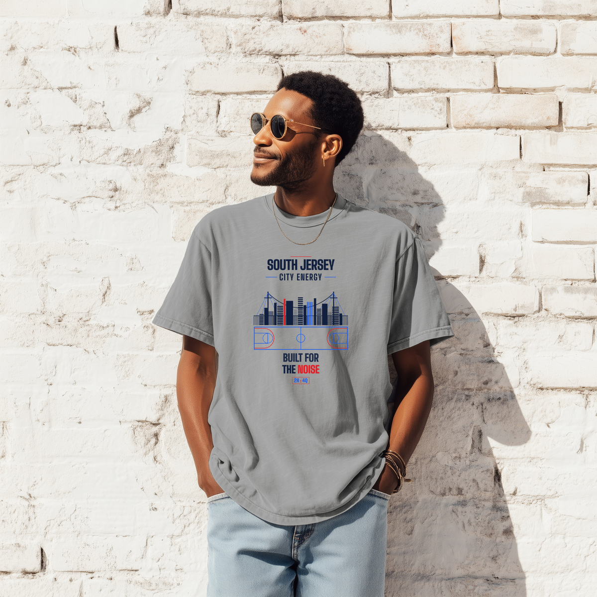 A man wearing a heather gray graphic t-shirt with a South Jersey city skyline print, standing against a white brick wall.