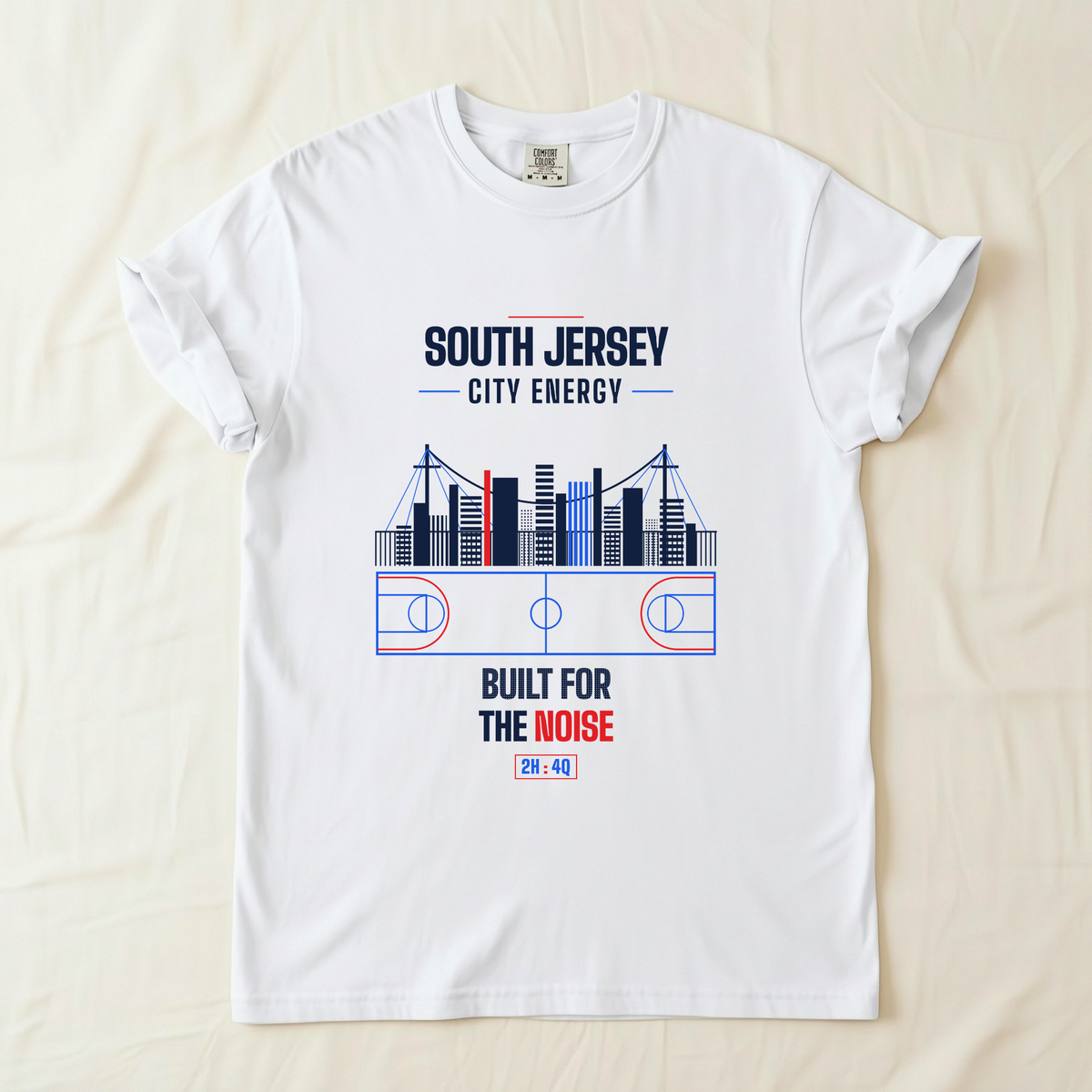 A white short-sleeve t-shirt laid flat on a light beige fabric background. The shirt features a graphic print of a basketball court and city skyline in blue, red, and navy blue, with the text 'SOUTH JERSEY CITY ENERGY' and 'BUILT FOR THE NOISE'.