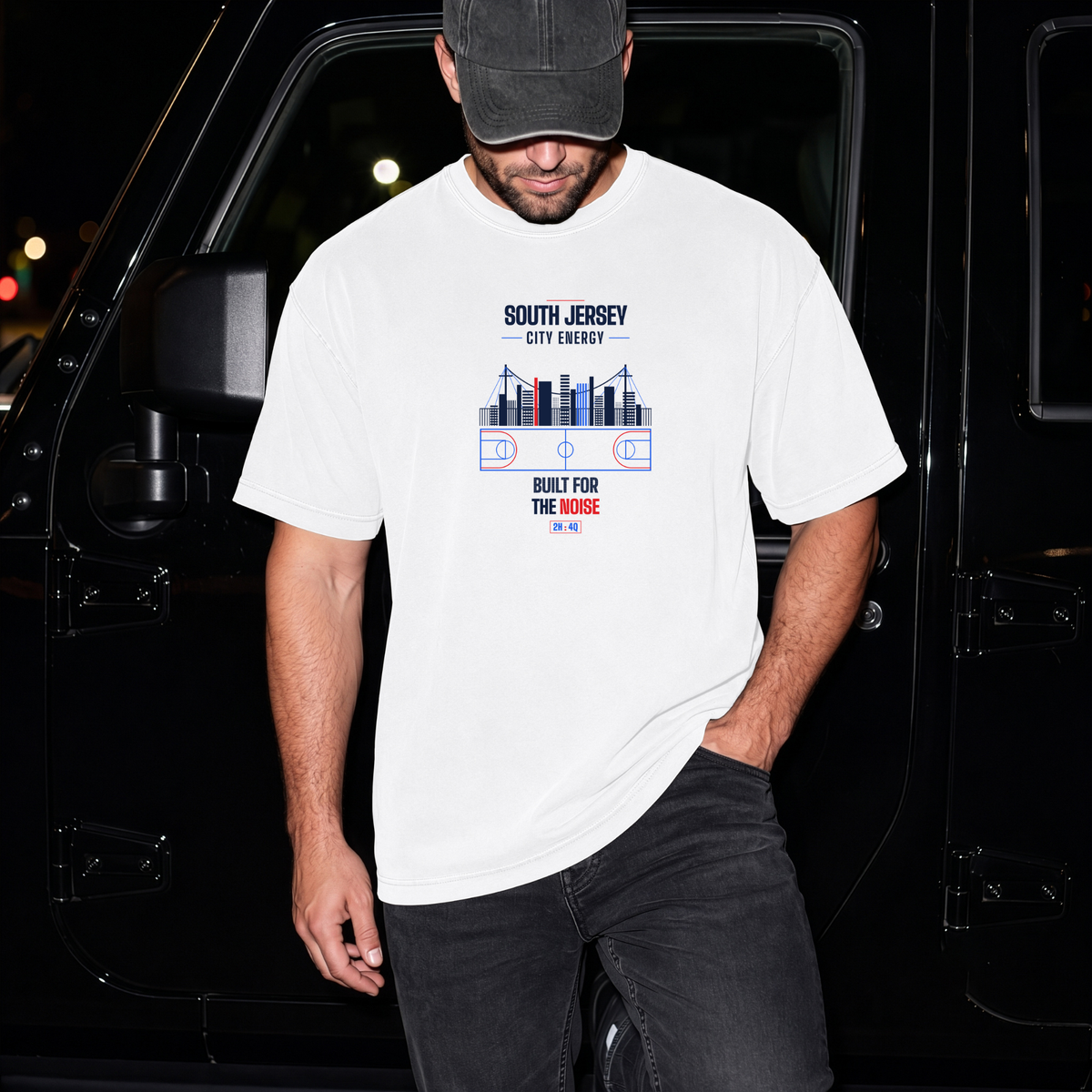 A man wearing a white graphic t-shirt with a 'South Jersey City Energy' design, paired with a black cap and dark jeans, standing in front of a black vehicle at night.