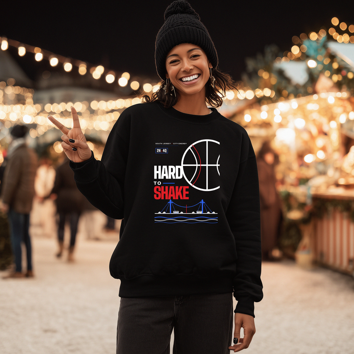A black woman wearing a black crewneck sweatshirt with a 'Hard to Shake' basketball graphic stands in a festive outdoor night market with warm string lights in the background.