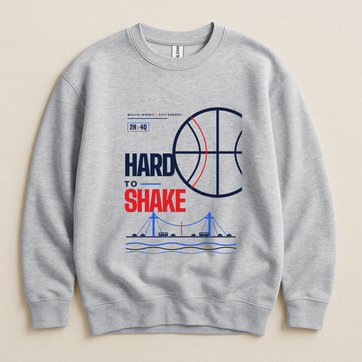 A light gray crewneck sweatshirt with a graphic print on the front featuring a basketball, a bridge, boats on water, and the text 'HARD TO SHAKE' in navy and red, displayed against a light beige background.