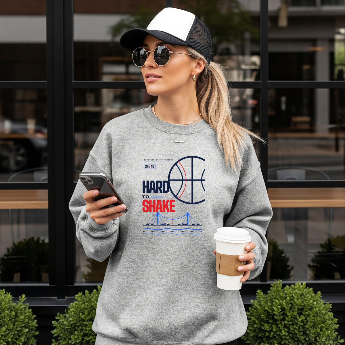 A woman wearing a light gray crewneck sweatshirt with a basketball-themed graphic that reads 'HARD TO SHAKE' stands in front of a storefront window, holding a phone and a coffee cup.