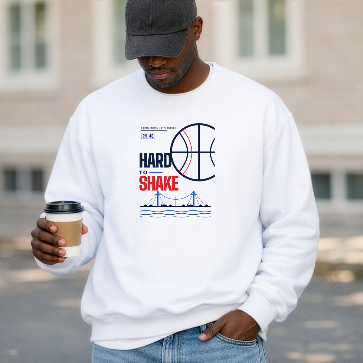 A black man wearing a white crewneck sweatshirt with a 'HARD TO SHAKE' basketball graphic, holding a coffee cup outdoors.