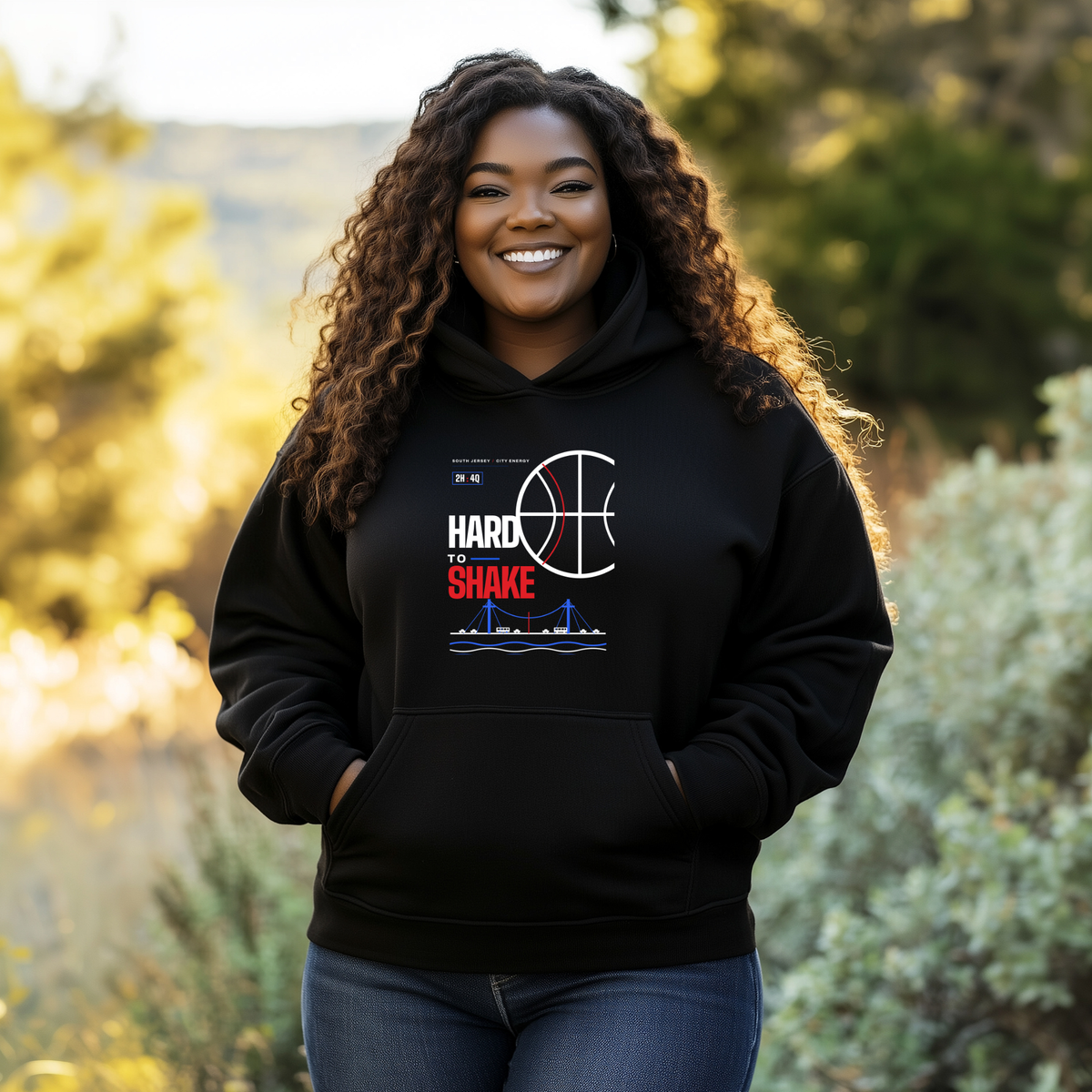 A smiling black woman stands outdoors wearing a black hoodie with a white, blue, and red graphic that reads 'HARD TO SHAKE' with a basketball and bridge design. The background shows blurred green foliage and warm sunlight.