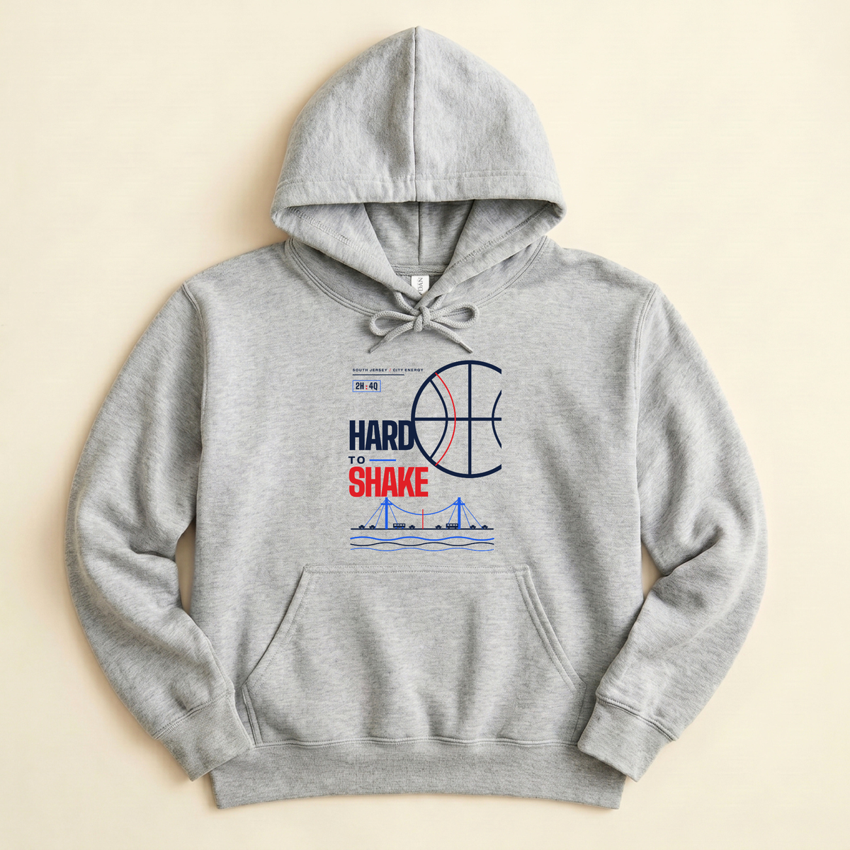 A light sport grey pullover hoodie with a graphic on the chest that reads 'HARD TO SHAKE' in blue and red, featuring a basketball and bridge illustration, laid flat on a light beige background.