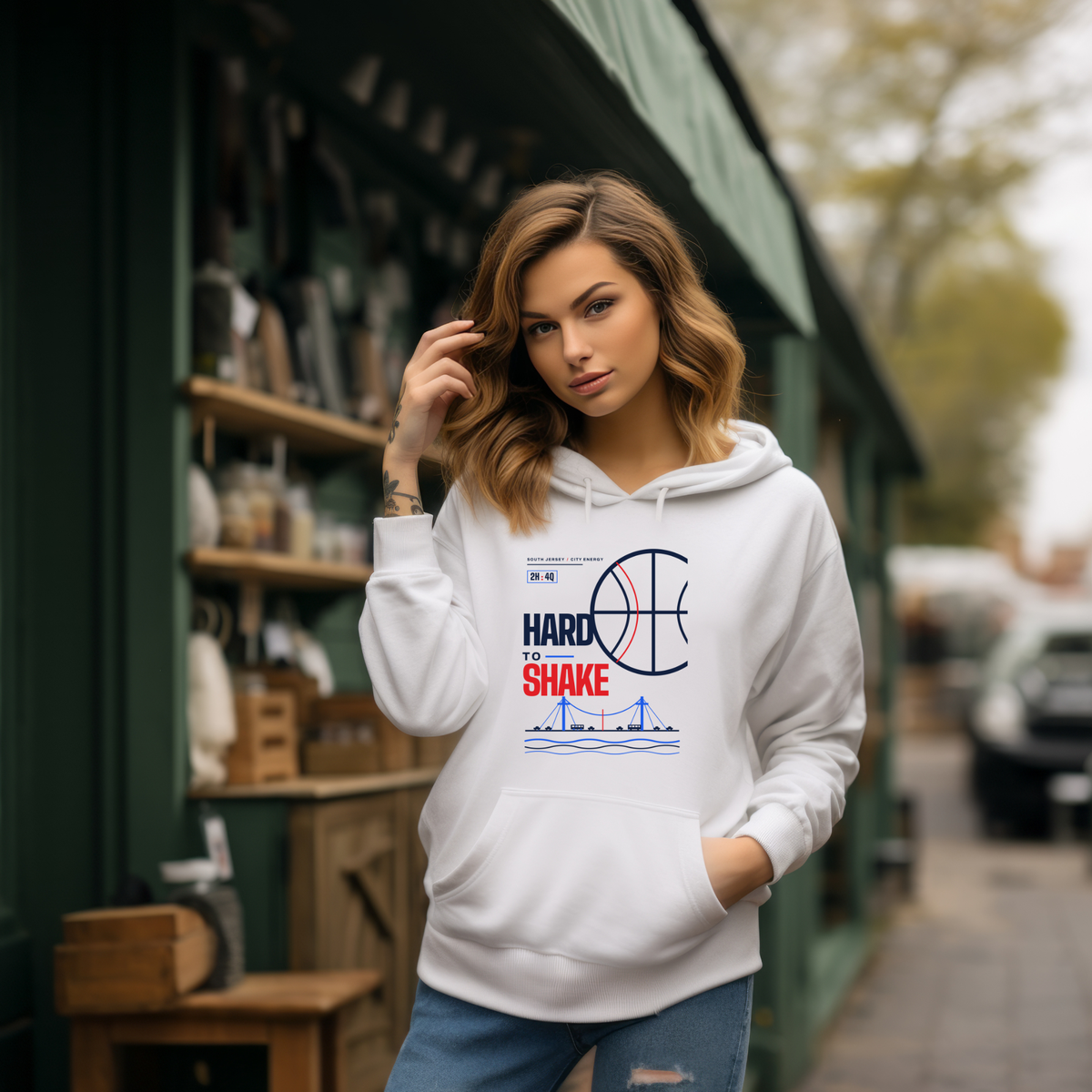 A woman wearing a white pullover hoodie with a colorful graphic print stands in front of a rustic outdoor shop. The hoodie features a basketball design and the text 'HARD AS A SHAKE'.