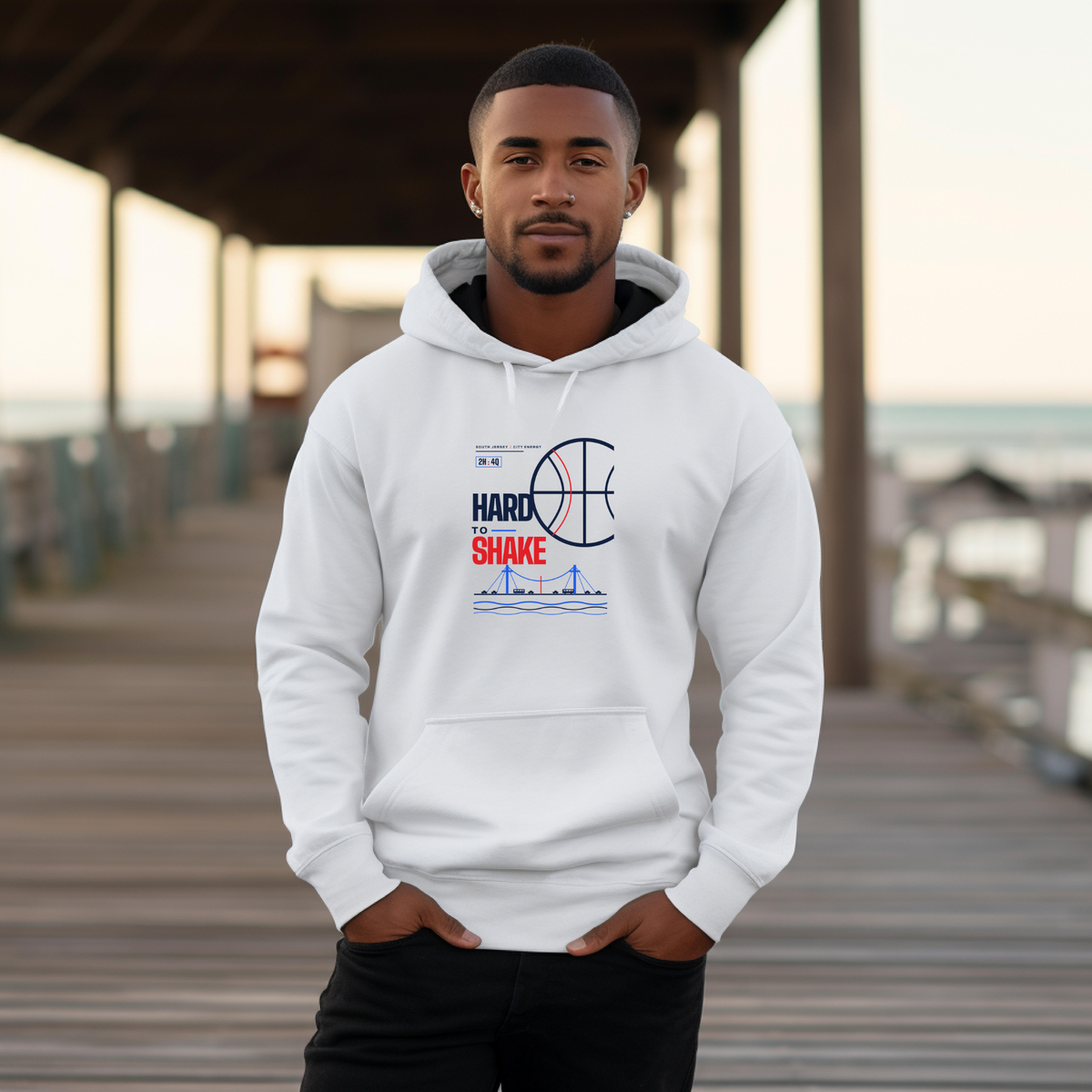 A black man wearing a white pullover hoodie with a red and blue 'HARD TO SHAKE' basketball graphic stands on a wooden boardwalk.