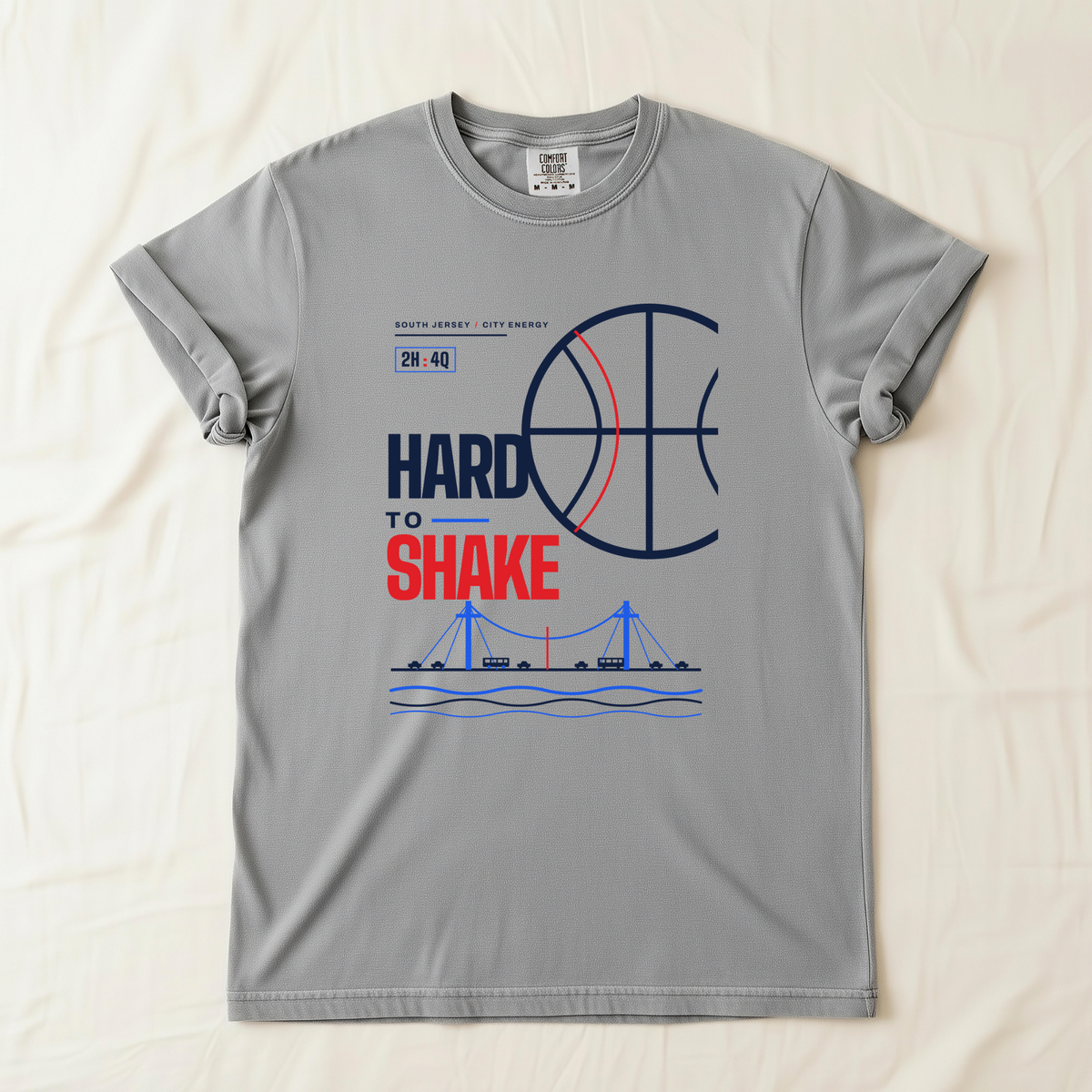 A gray short-sleeve t-shirt laid flat on a light beige fabric background. The shirt features a basketball-themed graphic with the text 'HARD TO SHAKE' in navy and red, a basketball design, and a blue bridge illustration below.