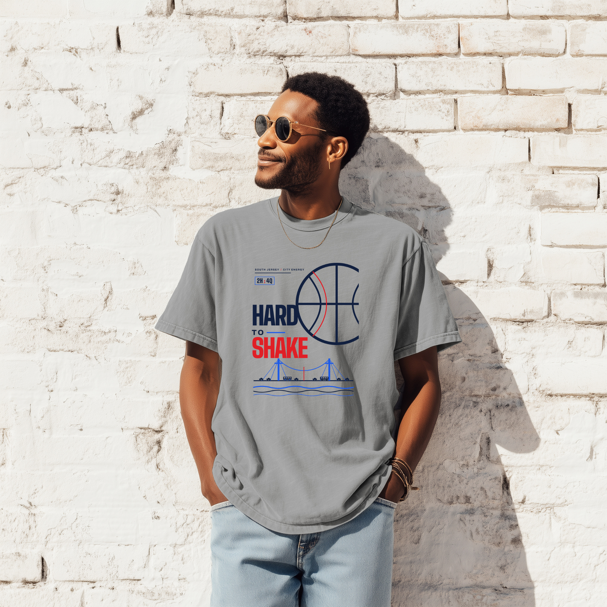 A black man wearing a gray graphic t-shirt with a basketball-themed print that reads 'HARD TO SHAKE' , paired with light blue jeans, standing against a white brick wall.
