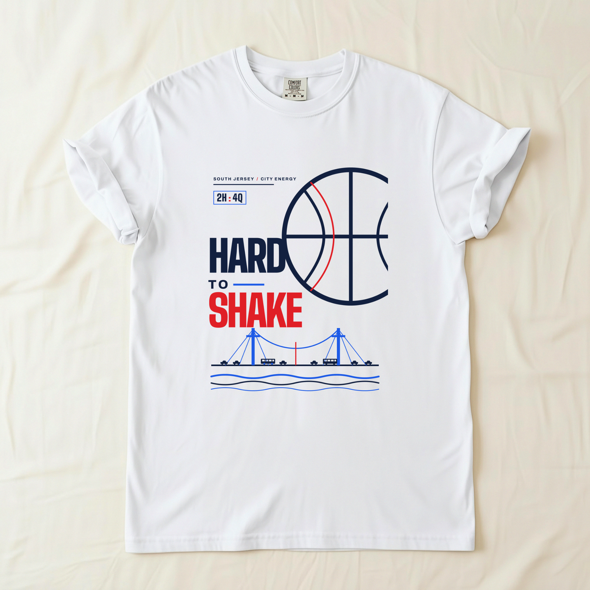 A white short-sleeve t-shirt laid flat on a light beige fabric background. The shirt features a graphic print with a basketball, the text 'HARD TO SHAKE' in navy and red, '2H:4Q' in a small box, and a blue bridge over water near the bottom.