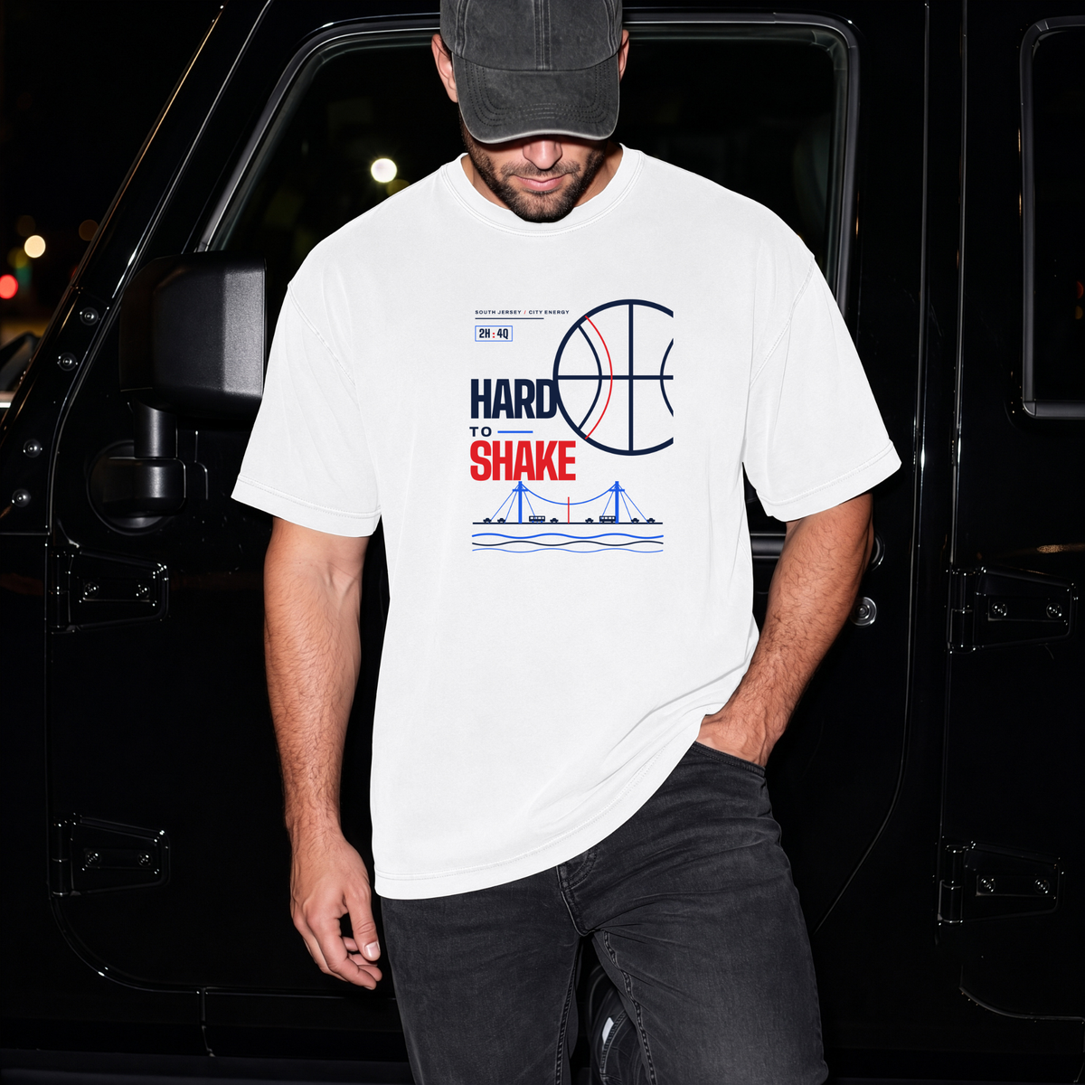 A man wearing a white graphic t-shirt with a basketball-themed 'Hard to Shake' design, paired with a dark cap and black jeans, standing in front of a black vehicle at night.