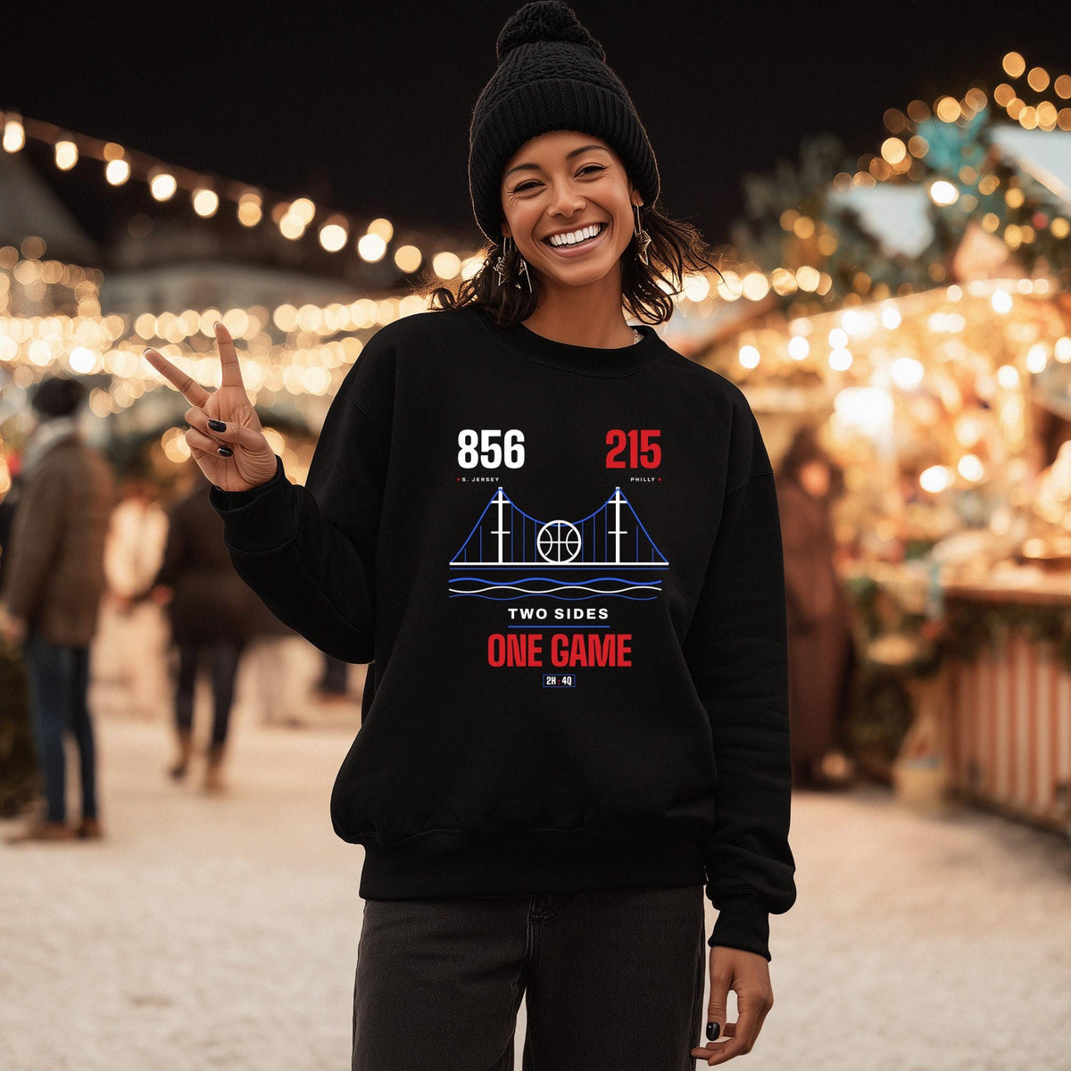 A black woman wearing a black crewneck sweatshirt with a graphic print stands in a festive outdoor night market. The sweatshirt features a white and blue bridge design with the text '856', '215' area codes, and 'TWO SIDES ONE GAME' graphic.