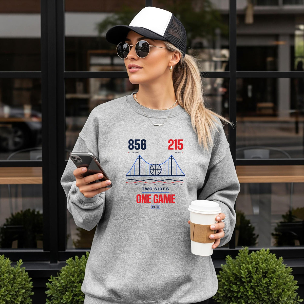 A woman wearing a light gray crewneck sweatshirt with a red and blue bridge graphic and the text '856 215 area codes, TWO SIDES ONE GAME'. She is holding a phone and a coffee cup, standing in front of a modern building with large black-framed windows.