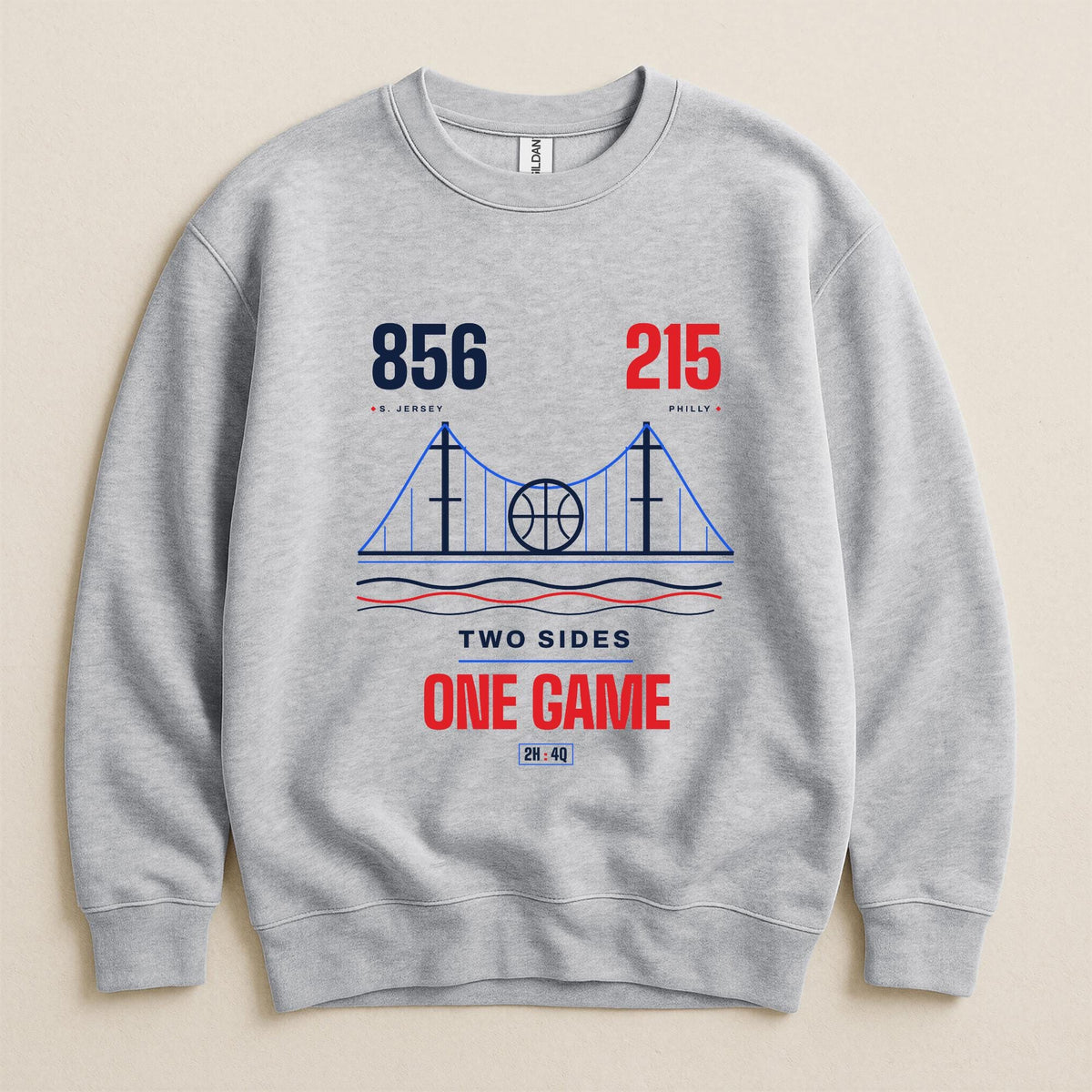 A light heather gray crewneck sweatshirt featuring a graphic print of the Golden Gate Bridge with a basketball in the center, flanked by the area codes 856 and 215, and the text 'TWO SIDES ONE GAME' in navy and red on a beige background.