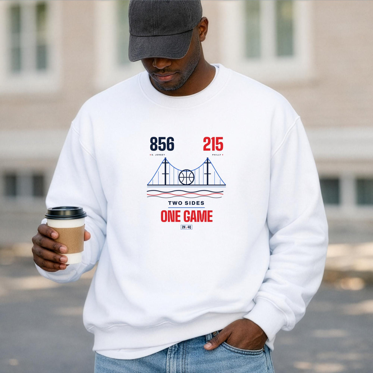 A black man wearing a white crewneck sweatshirt with a vintage-style sports graphic '856 and 215 area codes, TWO SIDES ONE GAME' on the chest, holding a coffee cup in front of a blurred outdoor background.