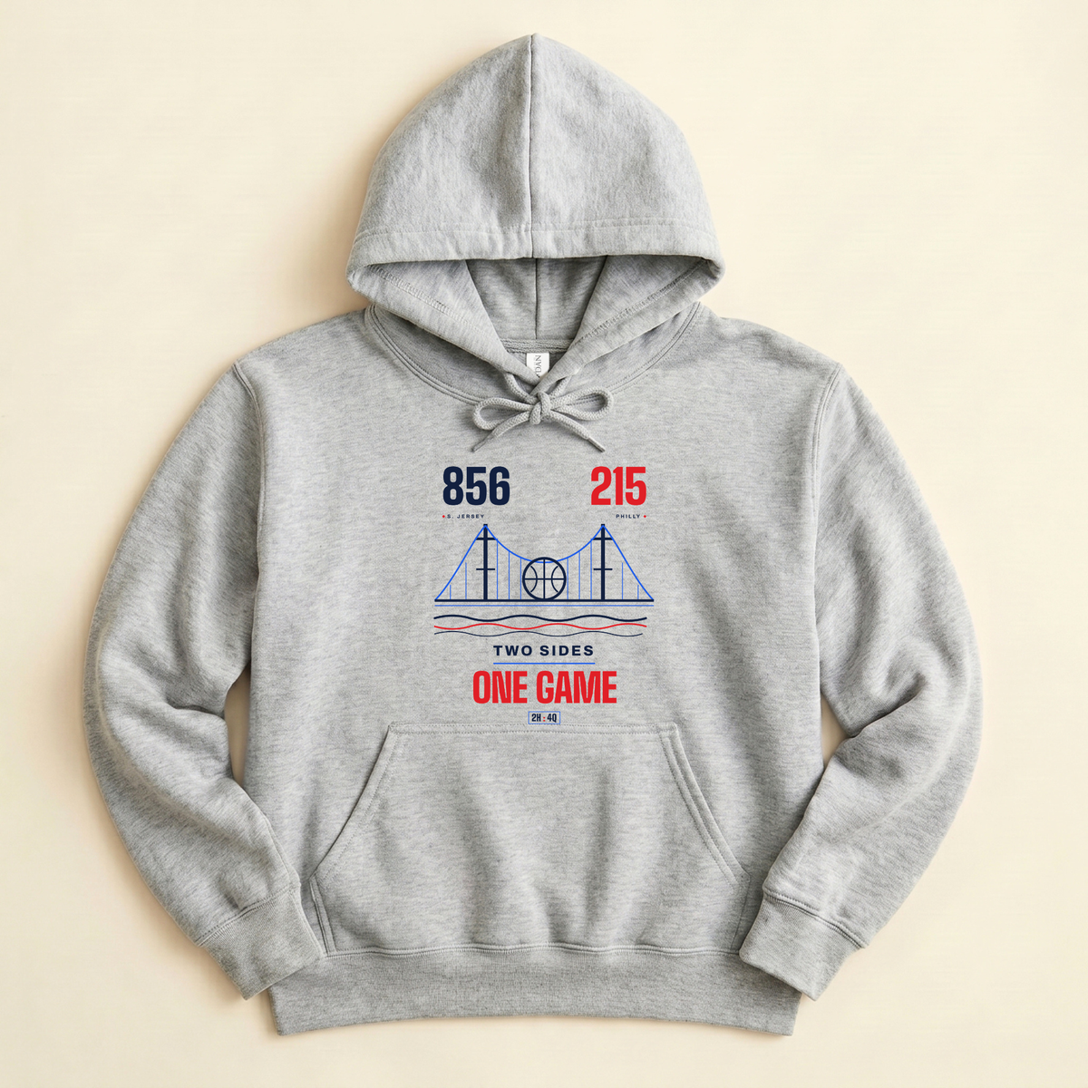 A light heather gray pullover hoodie with a vintage-style sports graphic on the chest, laid flat against a light beige background. The graphic features the numbers '856' and '215' area codes in navy blue and red, a blue bridge illustration, and the text 'TWO SIDES ONE GAME' in red and navy.