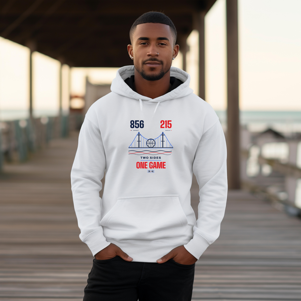 A black man wearing a white pullover hoodie with a red and blue, '856' and '215', 'TWO SIDES ONE GAME' sports graphic on the chest standing on a wooden boardwalk.