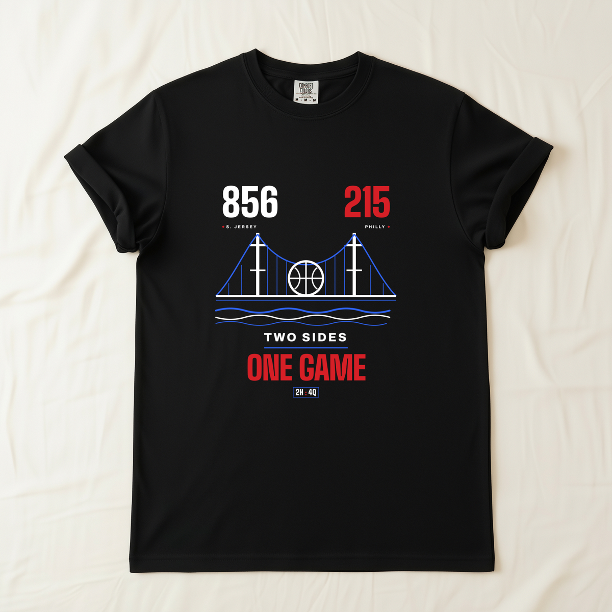 A black short-sleeve t-shirt laid flat on a light beige fabric background. The shirt features a graphic print of a bridge with a basketball in the center, flanked by the numbers '856' in white and '215' in red area codes, with the text 'TWO SIDES ONE GAME' in white and red below.