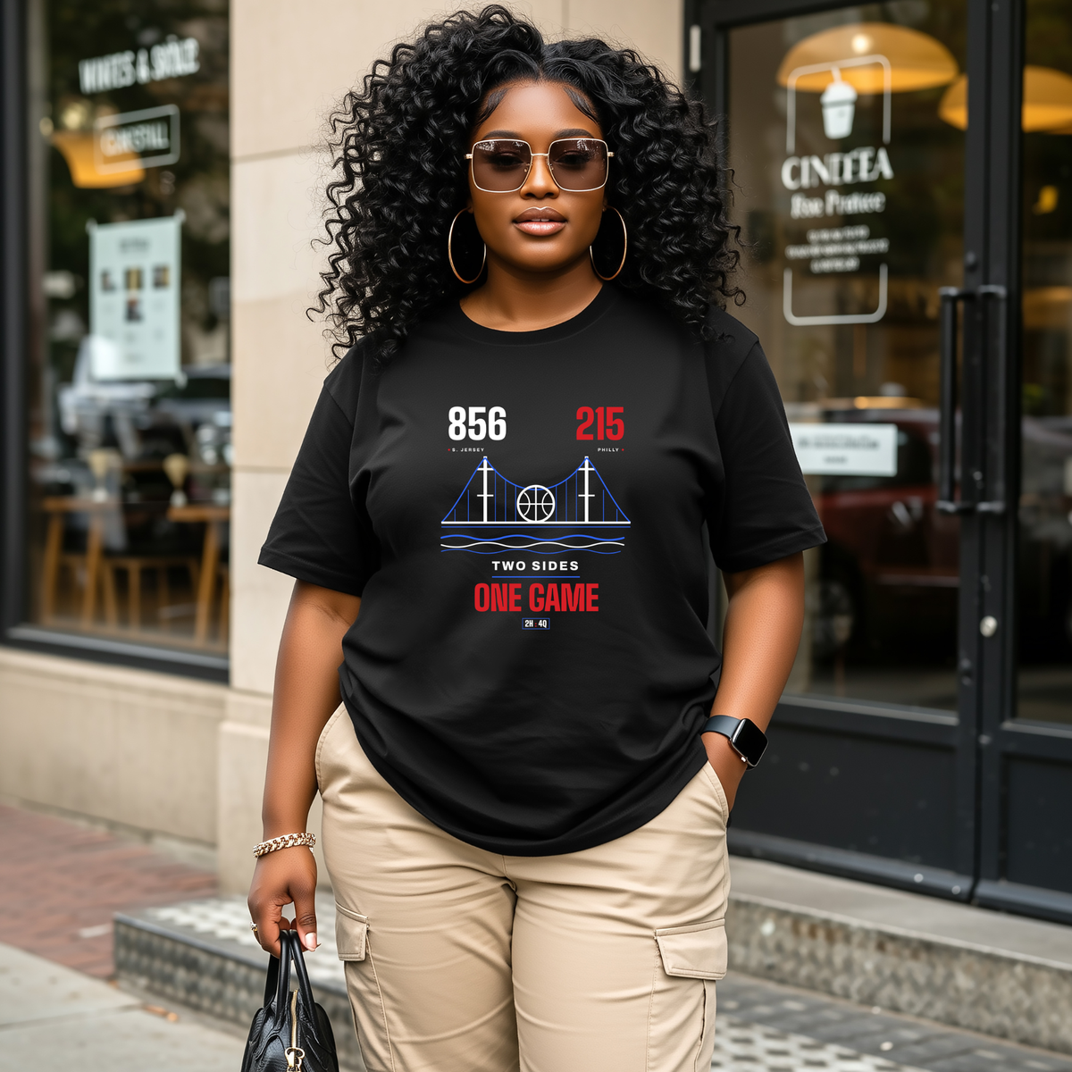 A black woman wearing a black graphic t-shirt with a red and blue '856' and '215', TWO SIDES ONE GAME, area code sports graphic print, styled with beige cargo pants and sunglasses, standing in front of a storefront.
