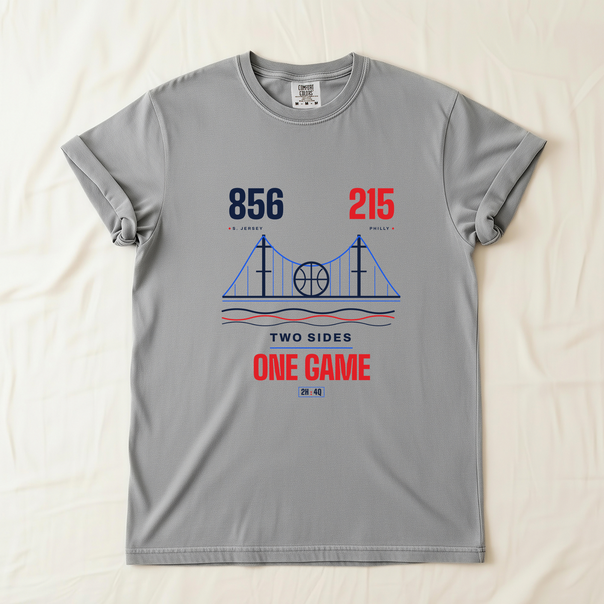 A light gray crewneck t-shirt laid flat on a cream-colored fabric background. The shirt features a graphic design of a basketball on a suspension bridge with the text '856', '215' area codes, 'TWO SIDES', and 'ONE GAME' in navy blue and red.