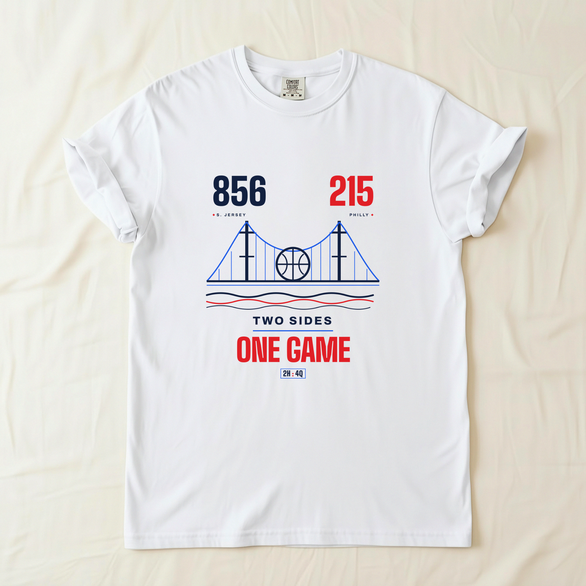 A white short-sleeve crewneck t-shirt laid flat on a light beige fabric background. The shirt features a graphic print of the a bridge with a basketball in the center, flanked by the numbers '856' area code in navy blue and '215' area code in red.