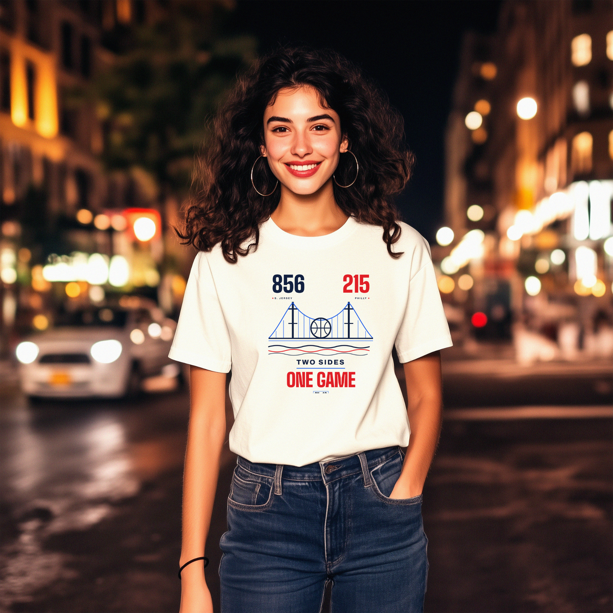 A smiling woman with curly dark hair stands on a blurred city street at night wearing a white graphic T-shirt and blue jeans. The shirt has a blue and red bridge graphic with the text '856, 215 area codes TWO SIDES ONE GAME'.