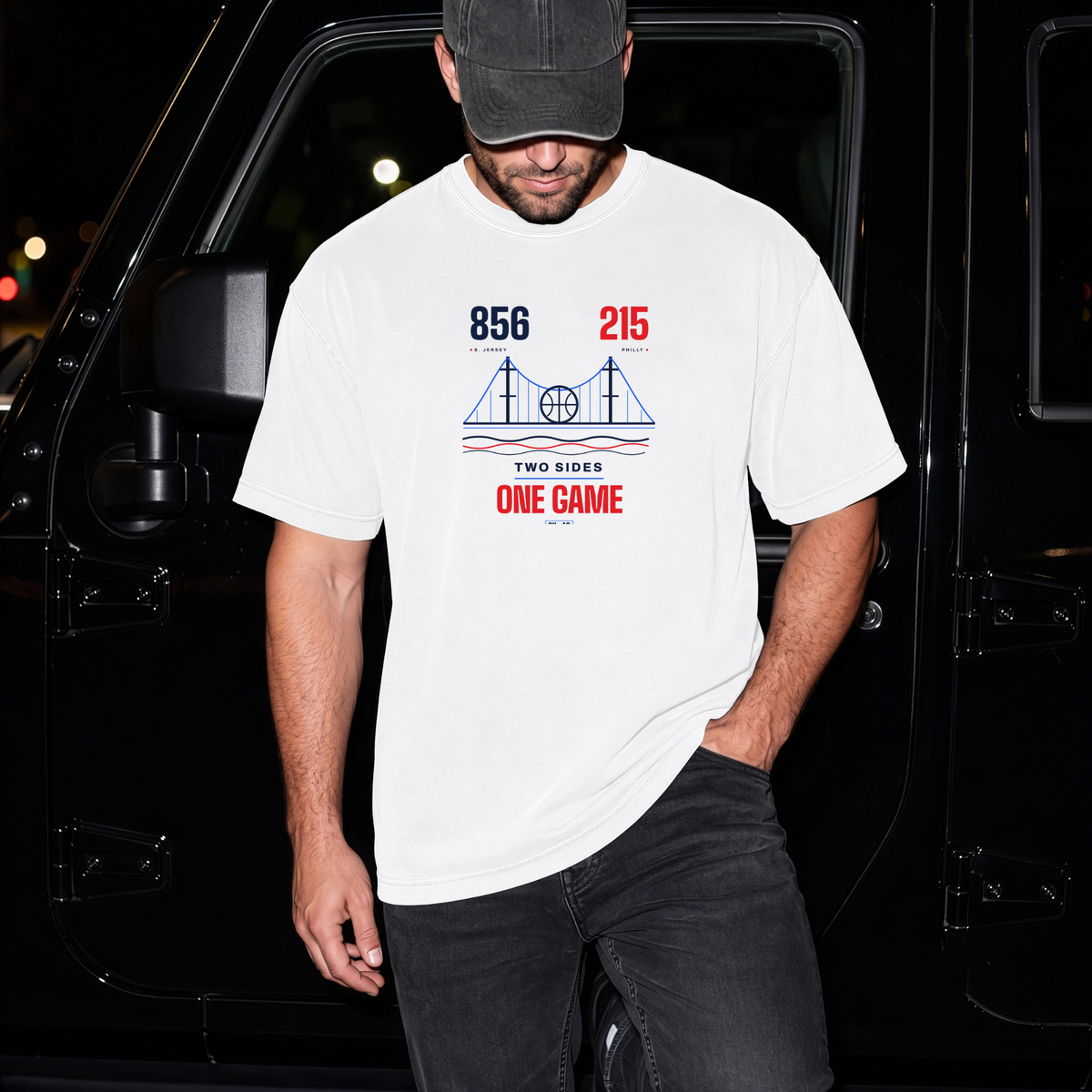 A man wearing a white graphic t-shirt with a red and blue 'Two Sides One Game' bridge design, paired with a dark cap and black jeans, standing in front of a black vehicle at night.